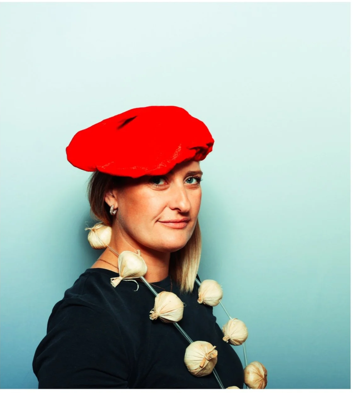 A woman wearing a red hat and a necklace made of garlic bulbs, smiling at the camera against a light blue background. Taken by Nook booth Photo Booth