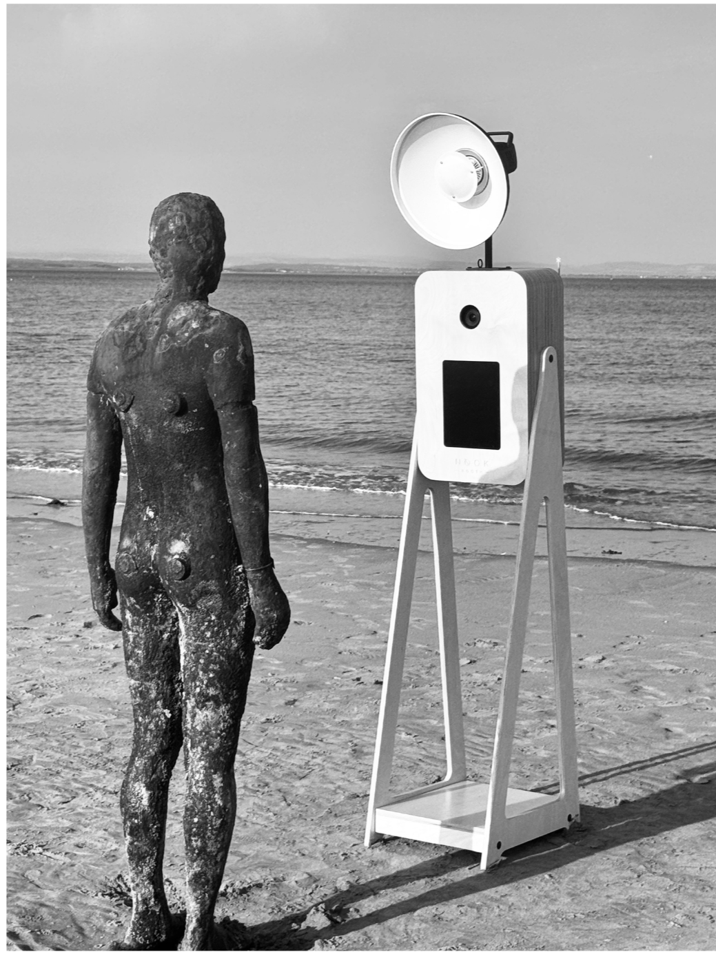 A weathered, life-size sculpture of a person standing on a sandy beach facing the ocean. Beside the sculpture is a modern photo booth or selfie station, equipped with a large light, a camera, and a screen, also positioned on the beach near the water.