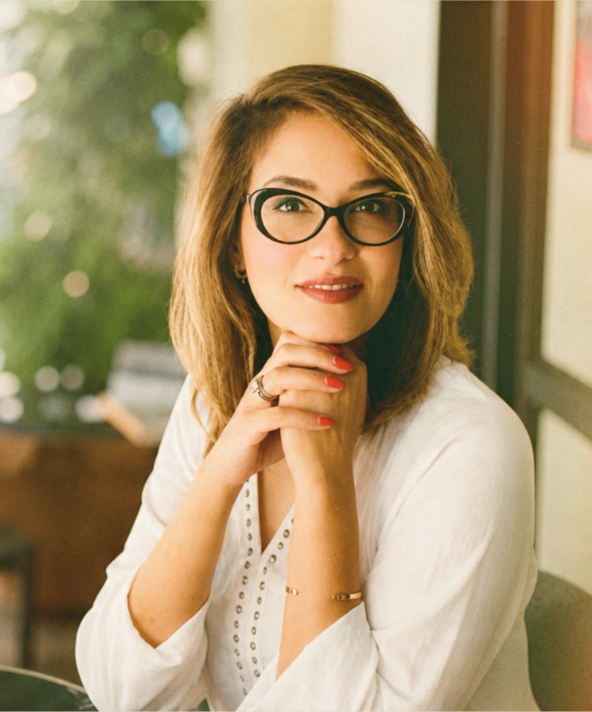 Close-up of a woman with shoulder-length light brown hair, wearing black-rimmed glasses, a white blouse, and jewelry, smiling with her hands clasped together under her chin, in a cozy indoor setting with blurred background.