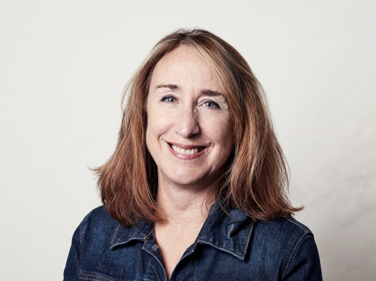 A woman with shoulder-length red hair, wearing a dark denim jacket, smiling at the camera against a plain light background.