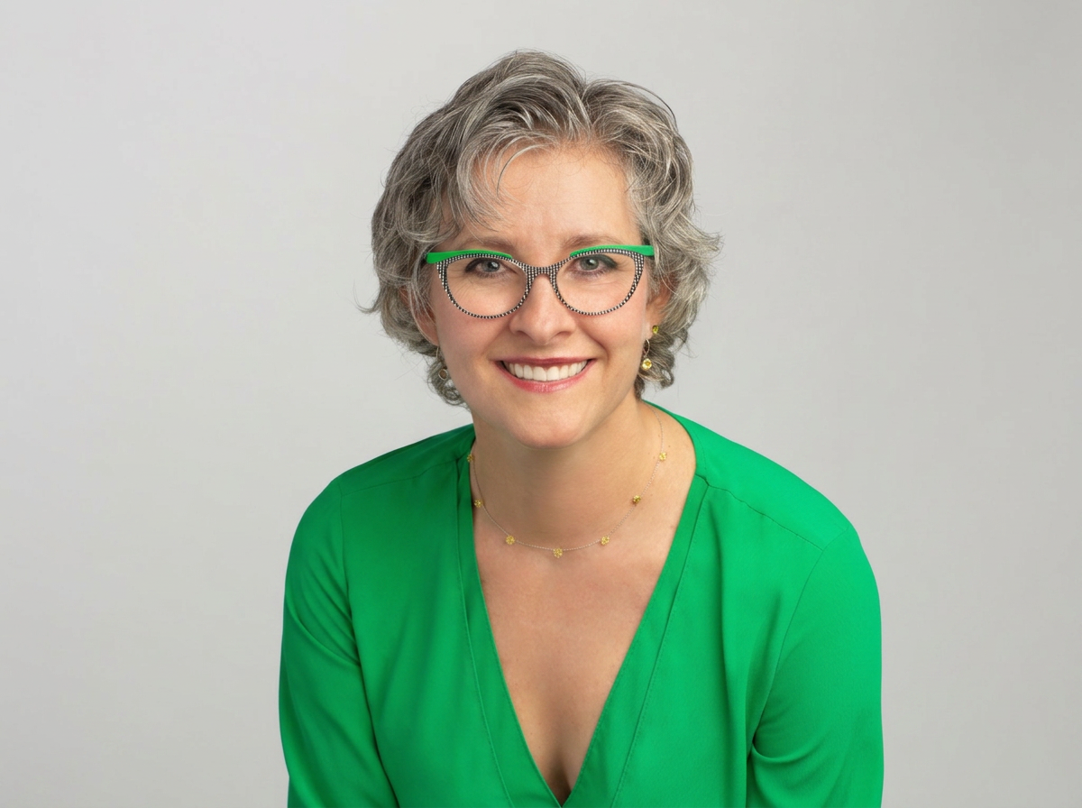 Portrait of a smiling woman with gray, curly hair wearing glasses with green accents, a green blouse, and a gold necklace, standing against a plain gray background.