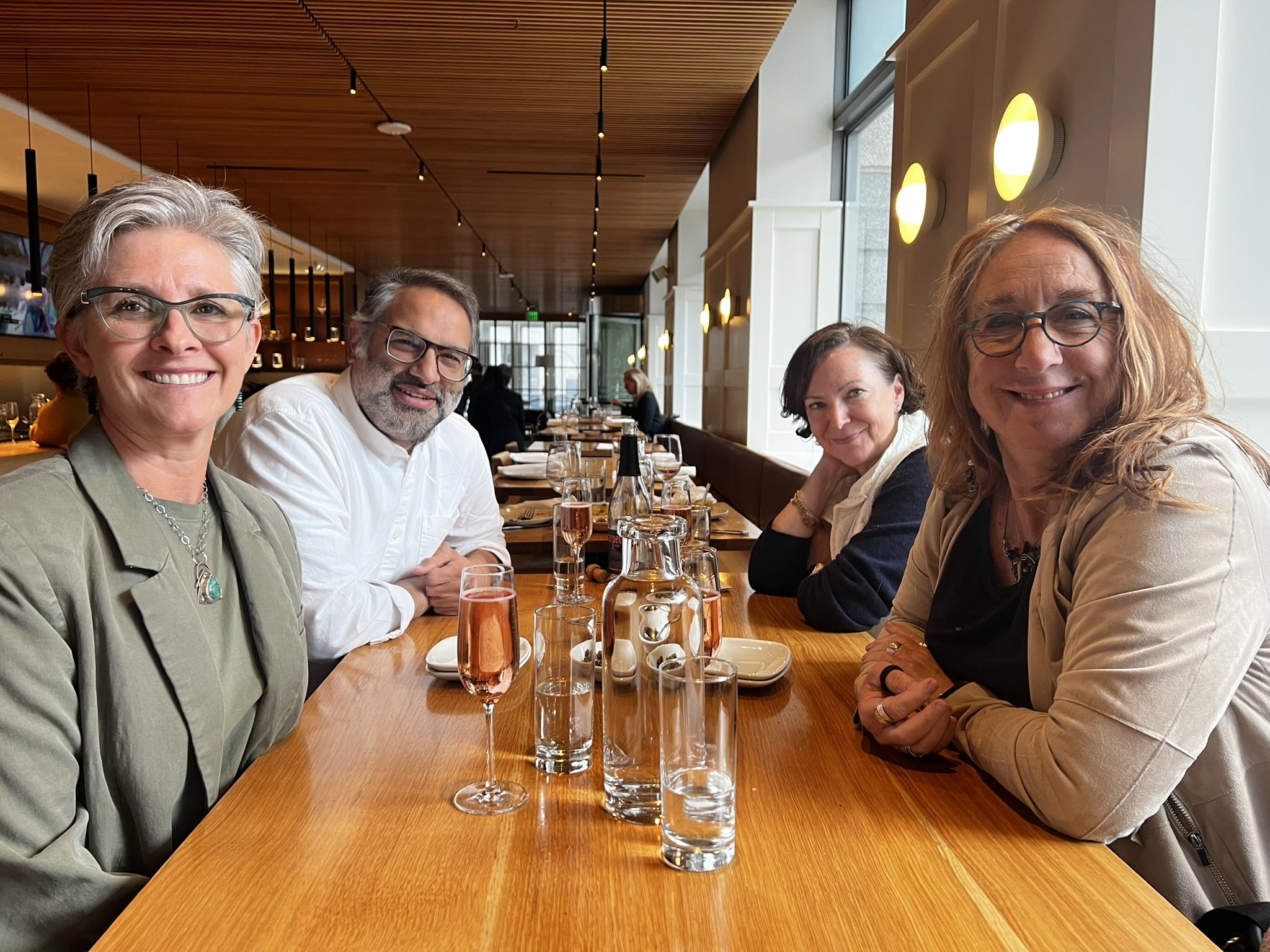 Four women and one man sitting at a long restaurant table with glasses of water and wine, smiling at the camera. The restaurant has a wooden ceiling and large windows.