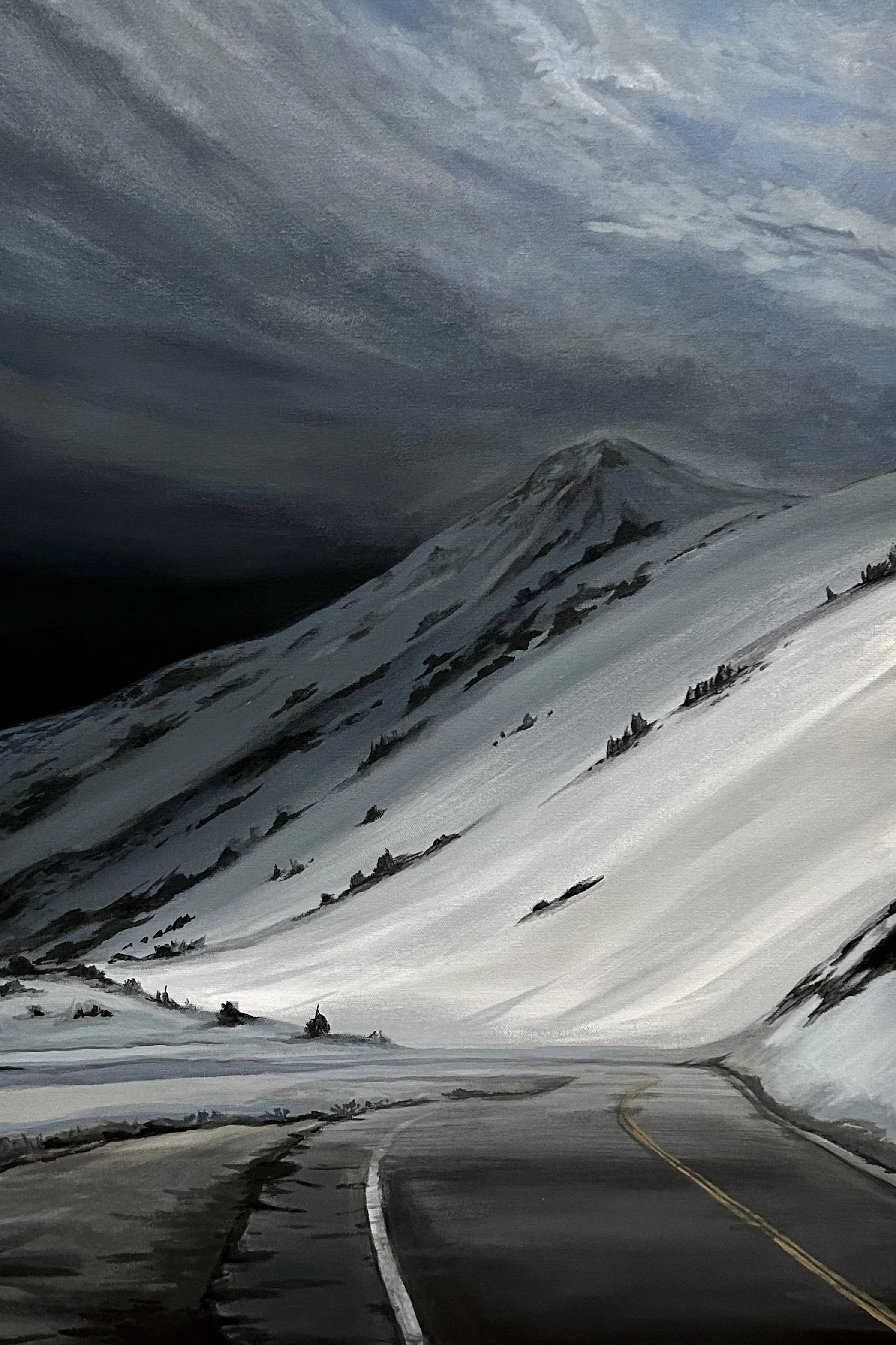 A winding mountain road covered in snow, with snow-covered slopes and a mountain in the background under a cloudy sky.