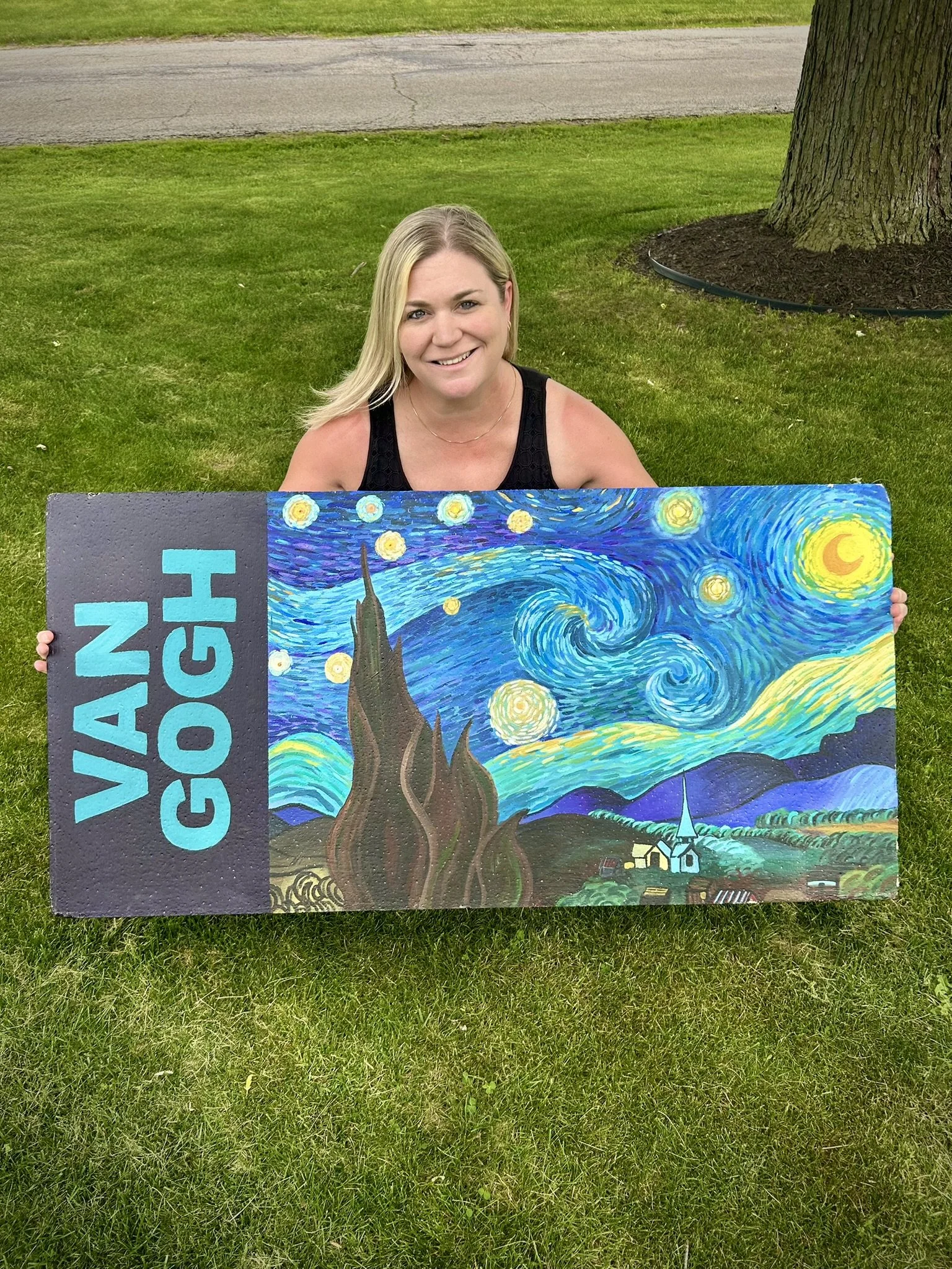 A smiling woman holding a colorful painting of Vincent van Gogh's Starry Night, with some text on it, outdoors on a grassy area next to a tree and a pavement.