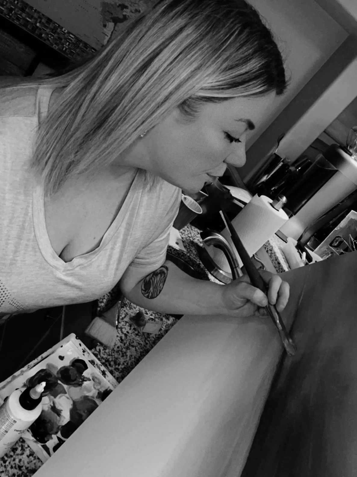 A woman with shoulder-length hair and a tattoo on her left arm, working on a piece of art or craft at a table in a well-lit room. Various art supplies and bottles are visible on the table around her.