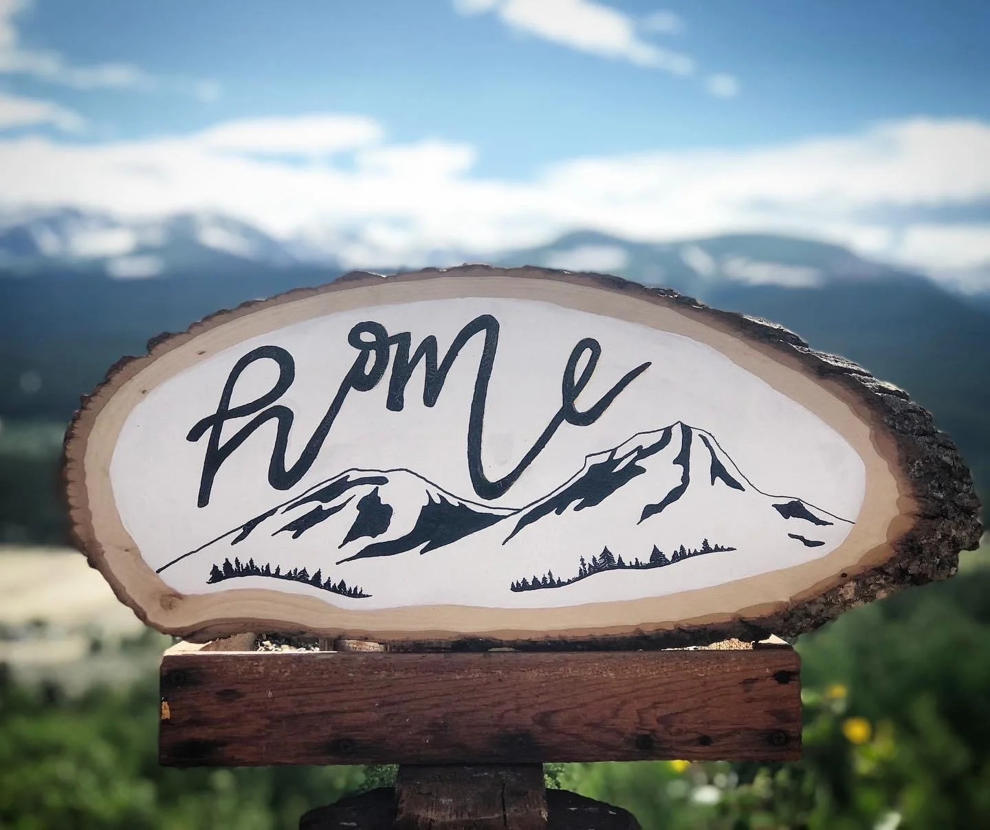 Wooden sign with the word "home" and mountain scenery, outdoors with mountains and blue sky in the background.