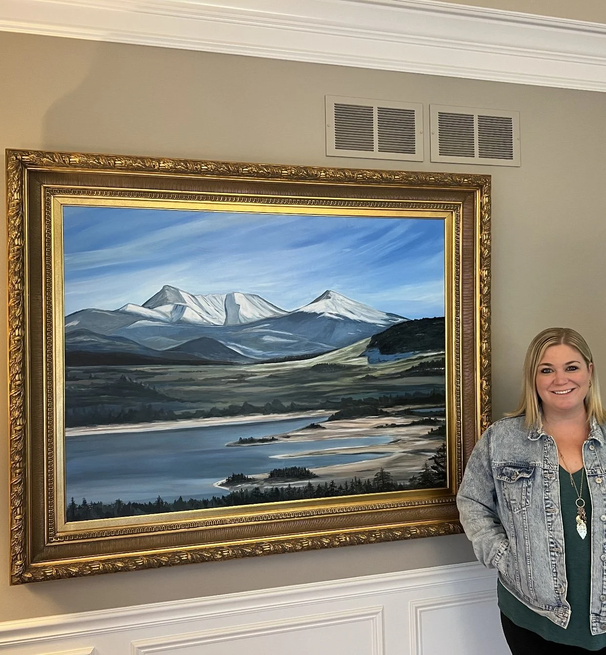 A woman standing next to a framed landscape painting of snow-capped mountains, a river, and green hills. The woman is smiling and wearing a denim jacket.