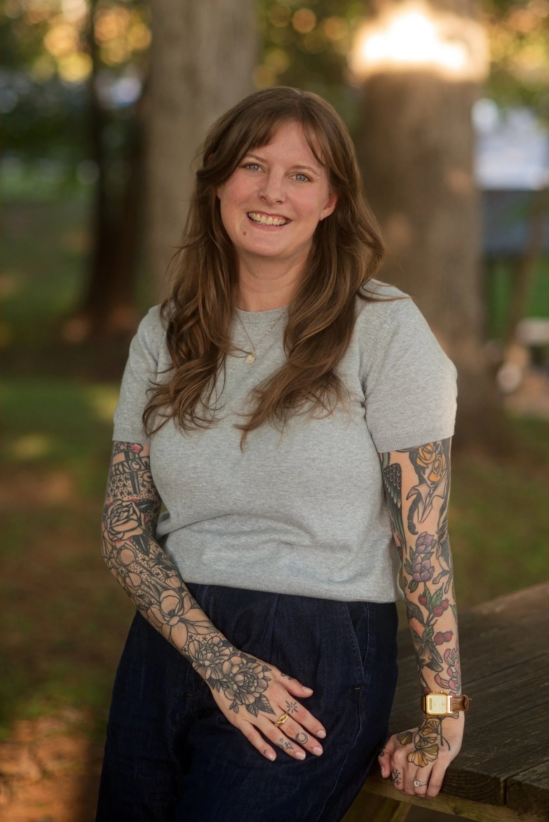 A woman with brown hair and tattoos on her arms, wearing a gray t-shirt and dark pants, standing outdoors by a wooden surface with trees and fall foliage in the background.