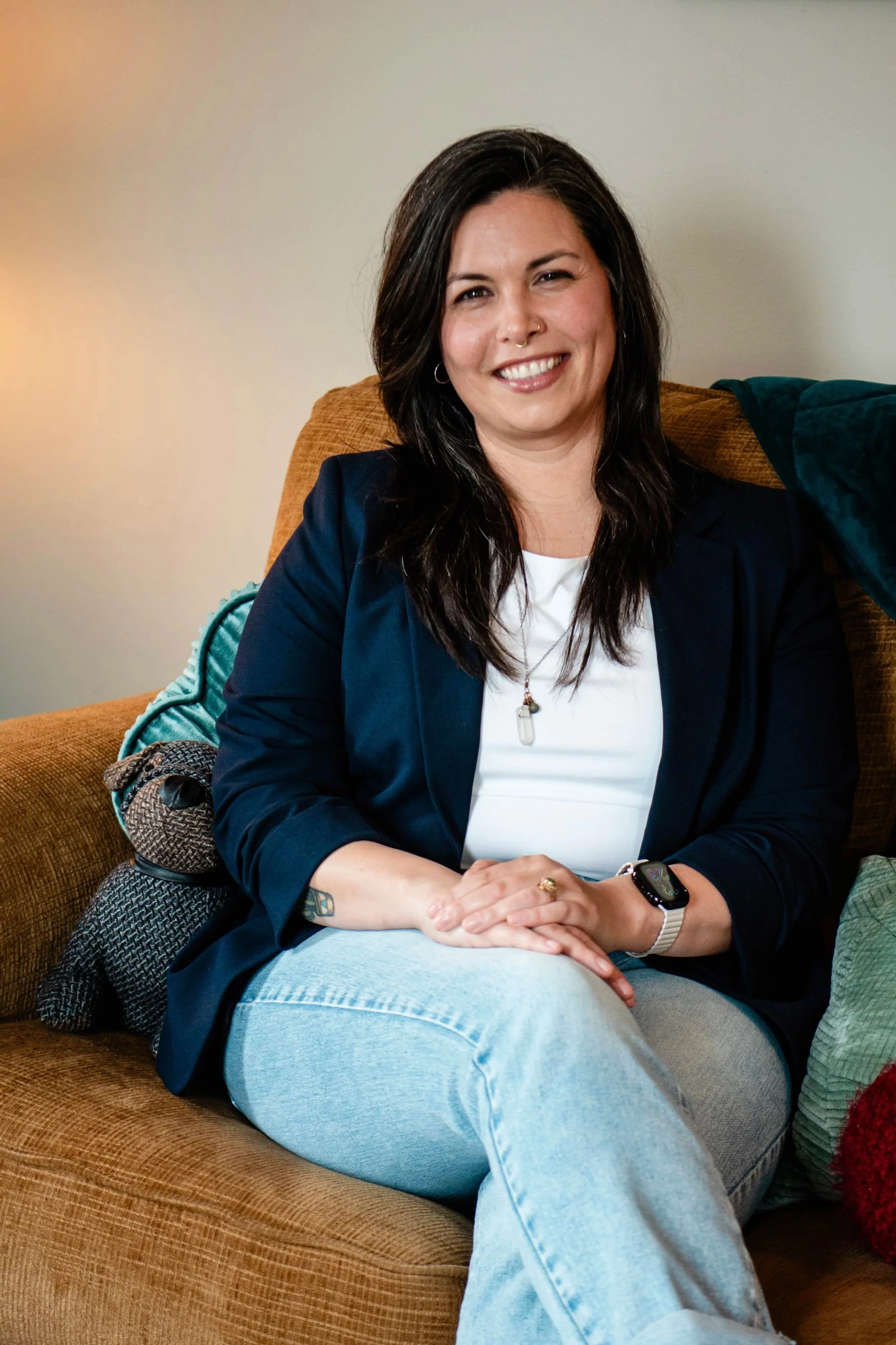 A woman with dark hair, wearing a navy blazer, white top, and light blue jeans, sitting on a brown couch, smiling, with a green pillow and a blanket next to her.