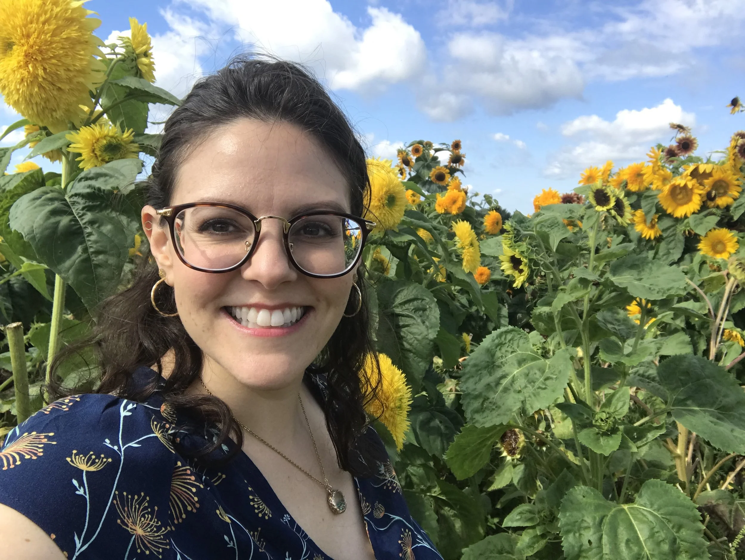 A woman with glasses and earrings smiling in a sunflower field on a partly cloudy day.