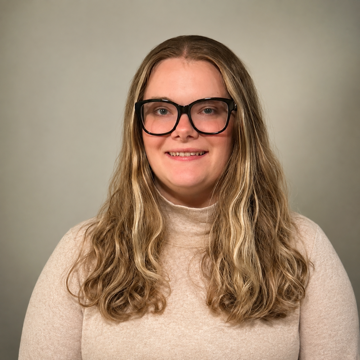 A woman with long, wavy blonde hair and black-rimmed glasses smiling in front of a plain light gray background.