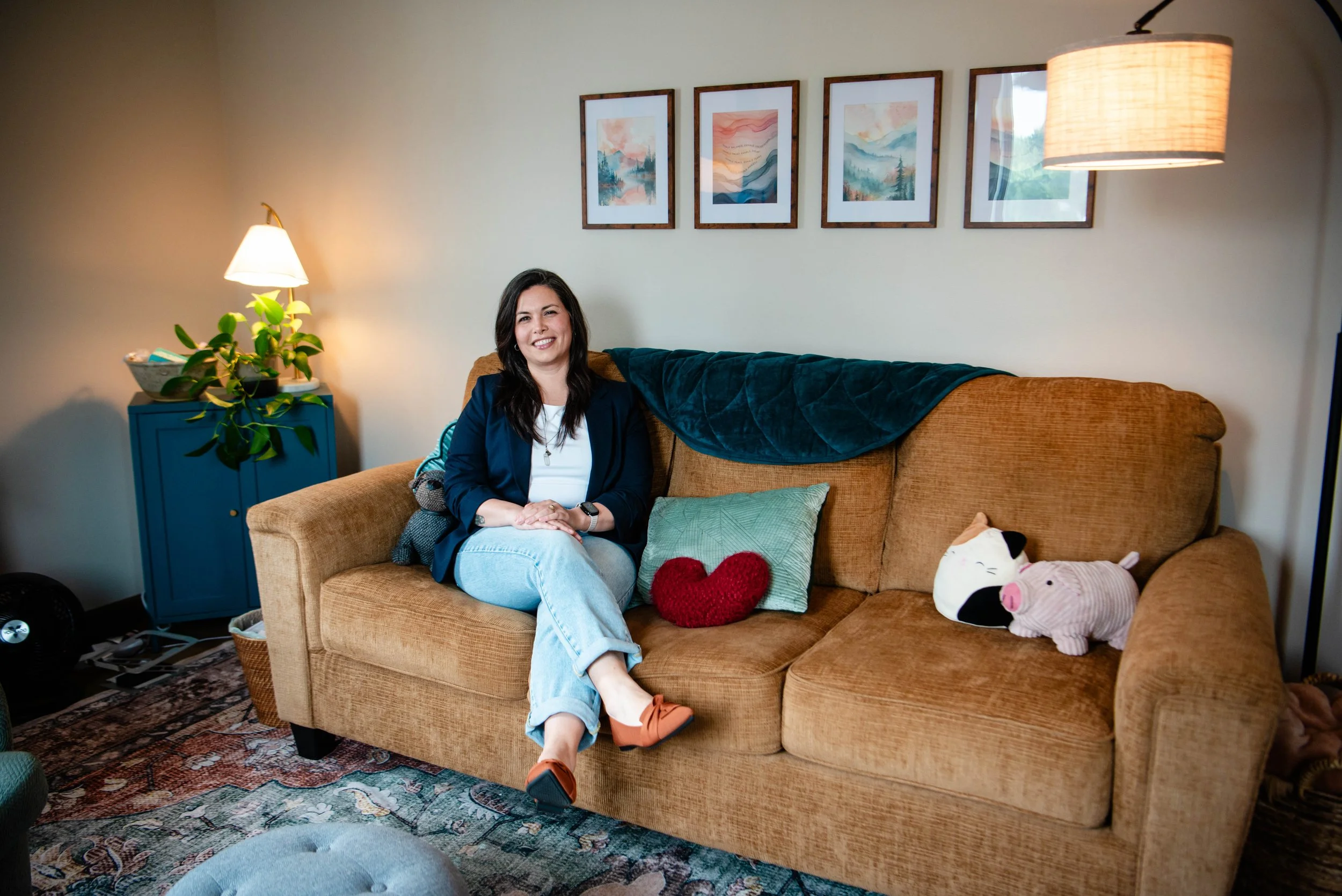 A woman sitting on a tan couch with a blue blanket draped over the backrest, smiling at the camera. The couch has decorative pillows, including a green pillow, a red hearts-shaped pillow, and a plush pig toy. Behind her, there's a wall with four framed landscape paintings. To the left, there's a blue side table with a plant and a lamp, and on the right, a standing lamp. The floor has a patterned rug.
