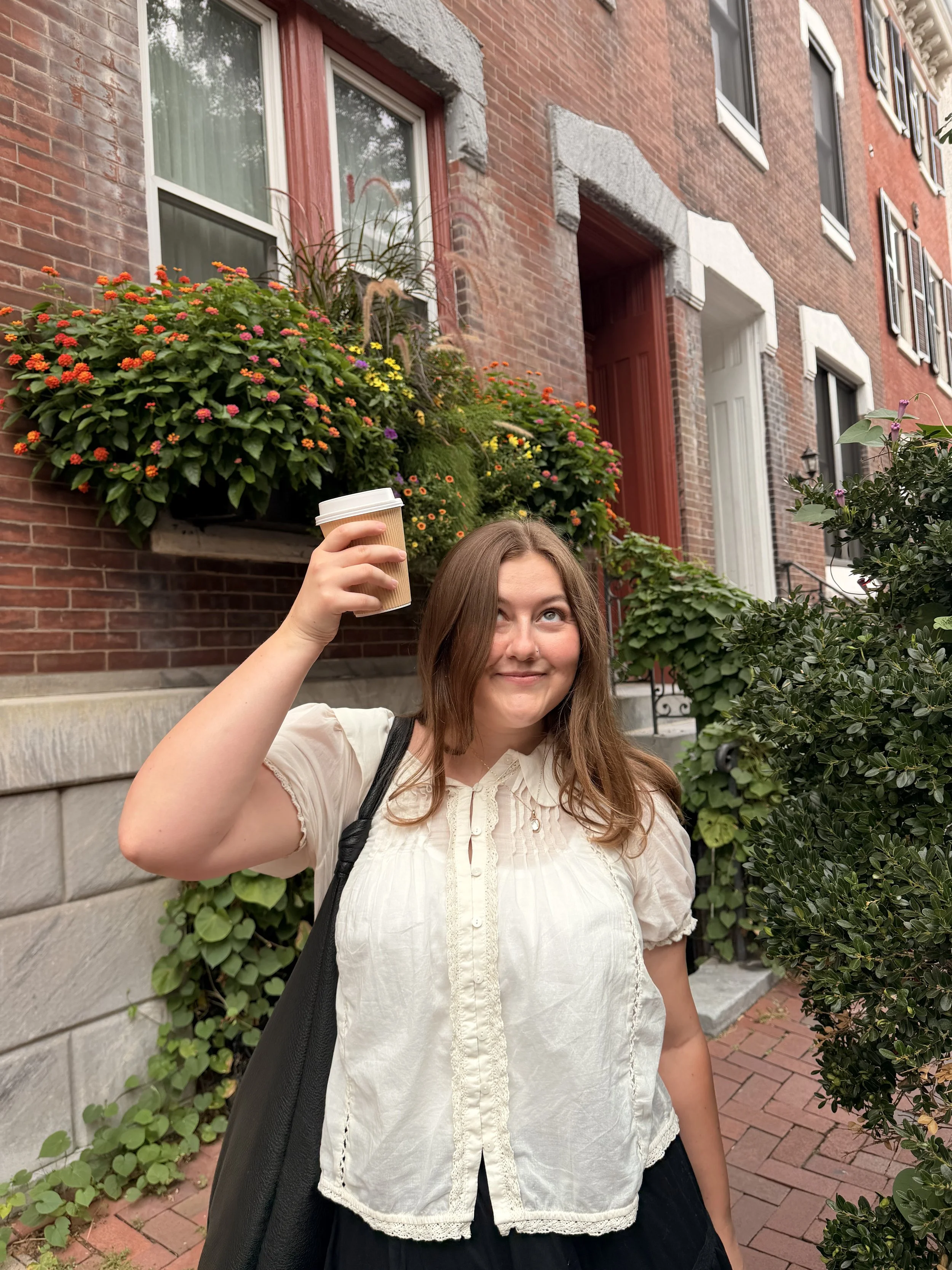 A smiling woman with long brown hair holding a disposable coffee cup, standing outdoors in front of a red brick building with windows and lush green plants.