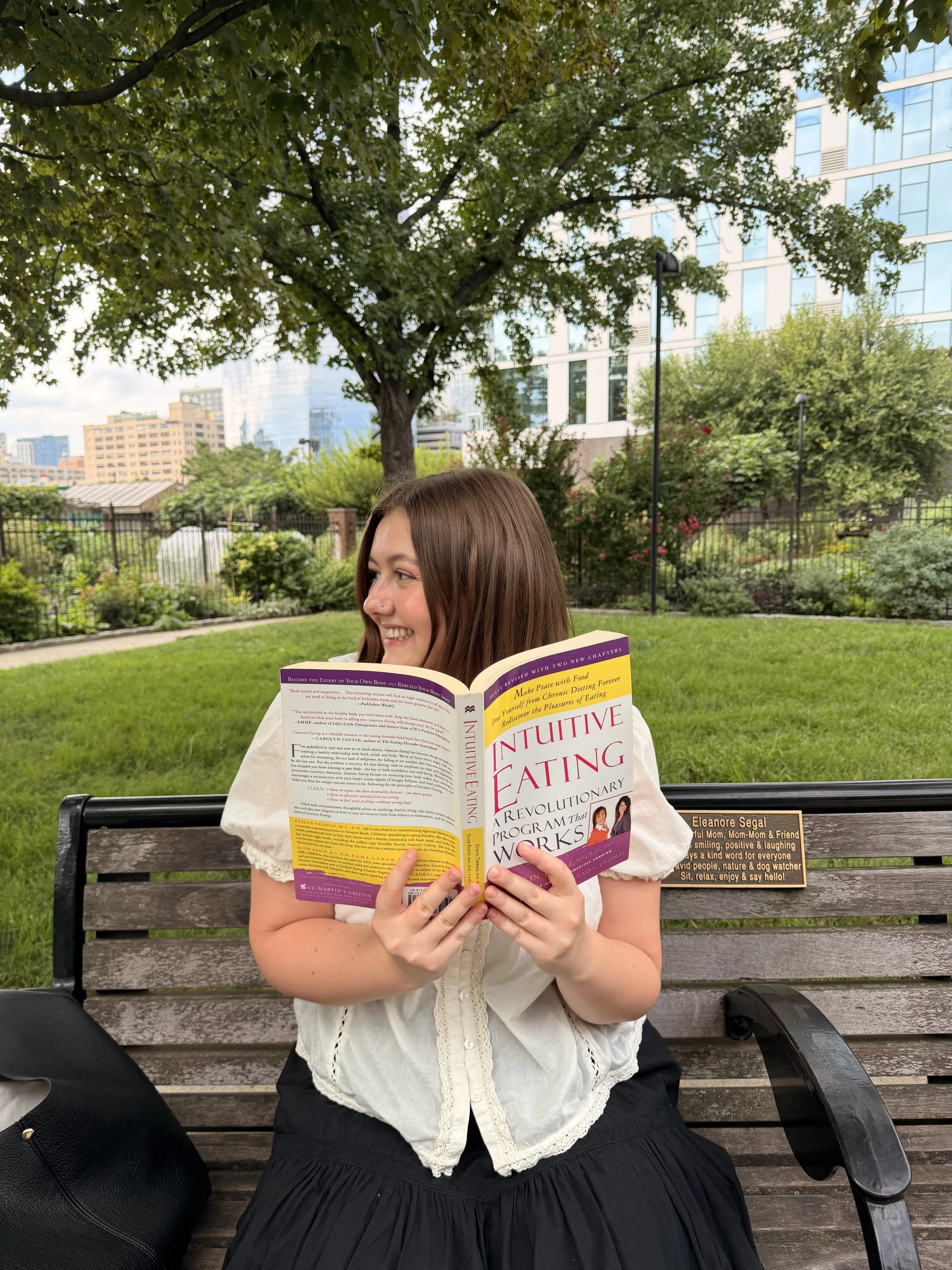 A woman sitting on a park bench reading a book titled 'Intuitive Eating' in a city park with trees and buildings in the background.