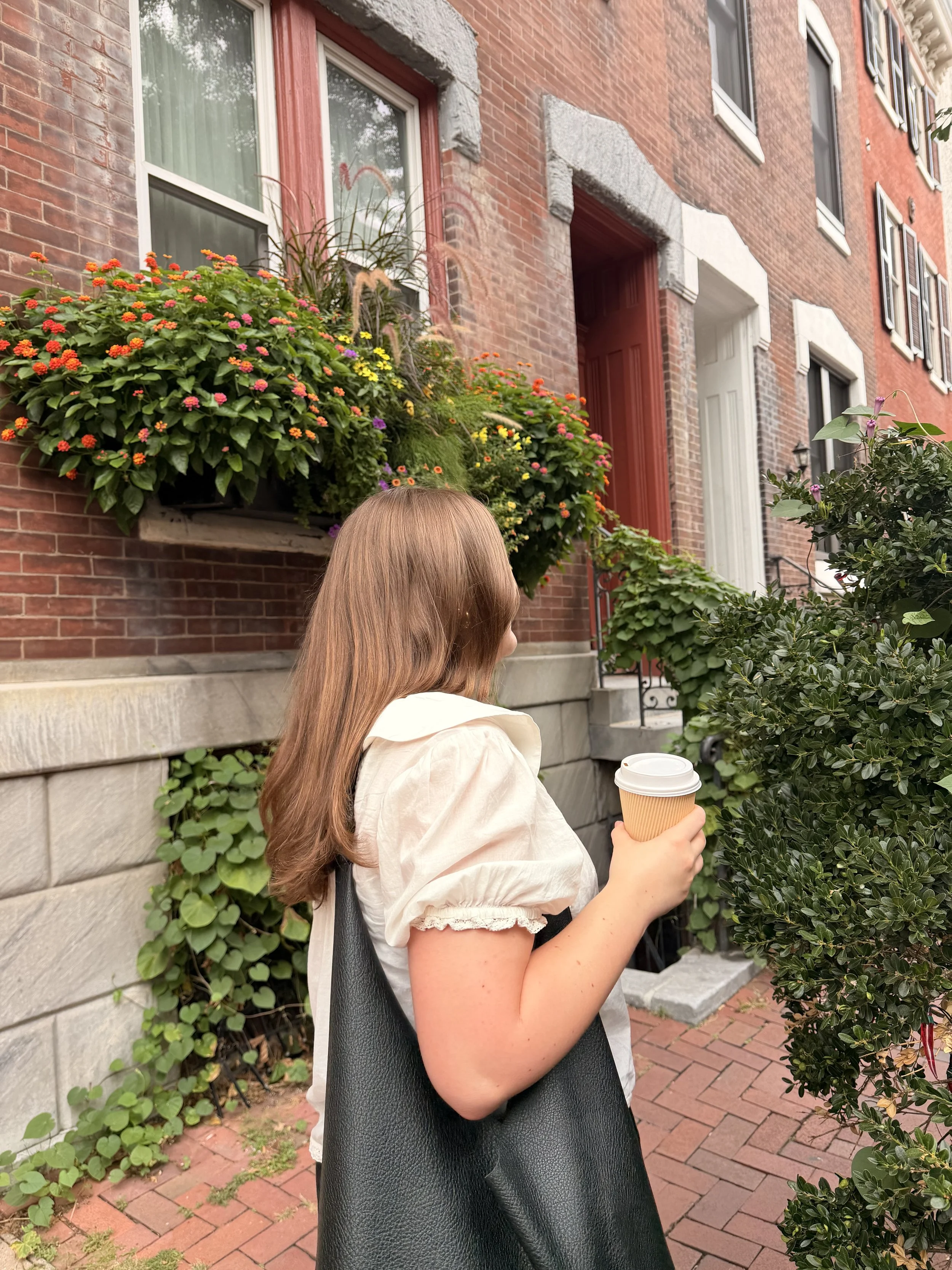 A woman with long brown hair holding a coffee cup in front of a lush green hedge and a brick house with flower boxes under the windows.