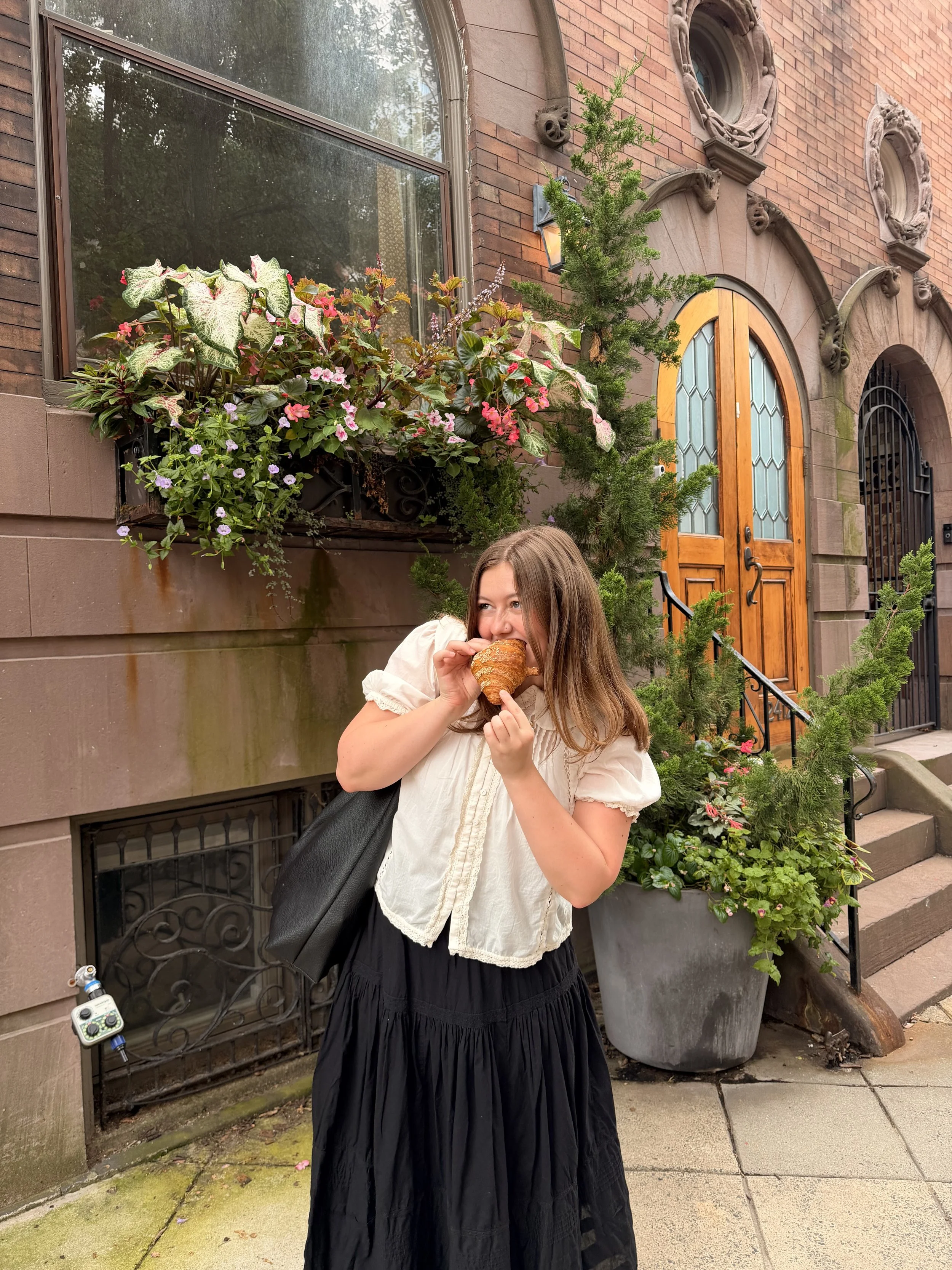 A woman with long brown hair wearing a white blouse and black skirt is standing outside in front of a brick building. She is eating a croissant and carrying a black shoulder bag. There are plants and flowers in large pots behind her, and a wooden door and large window on the building.