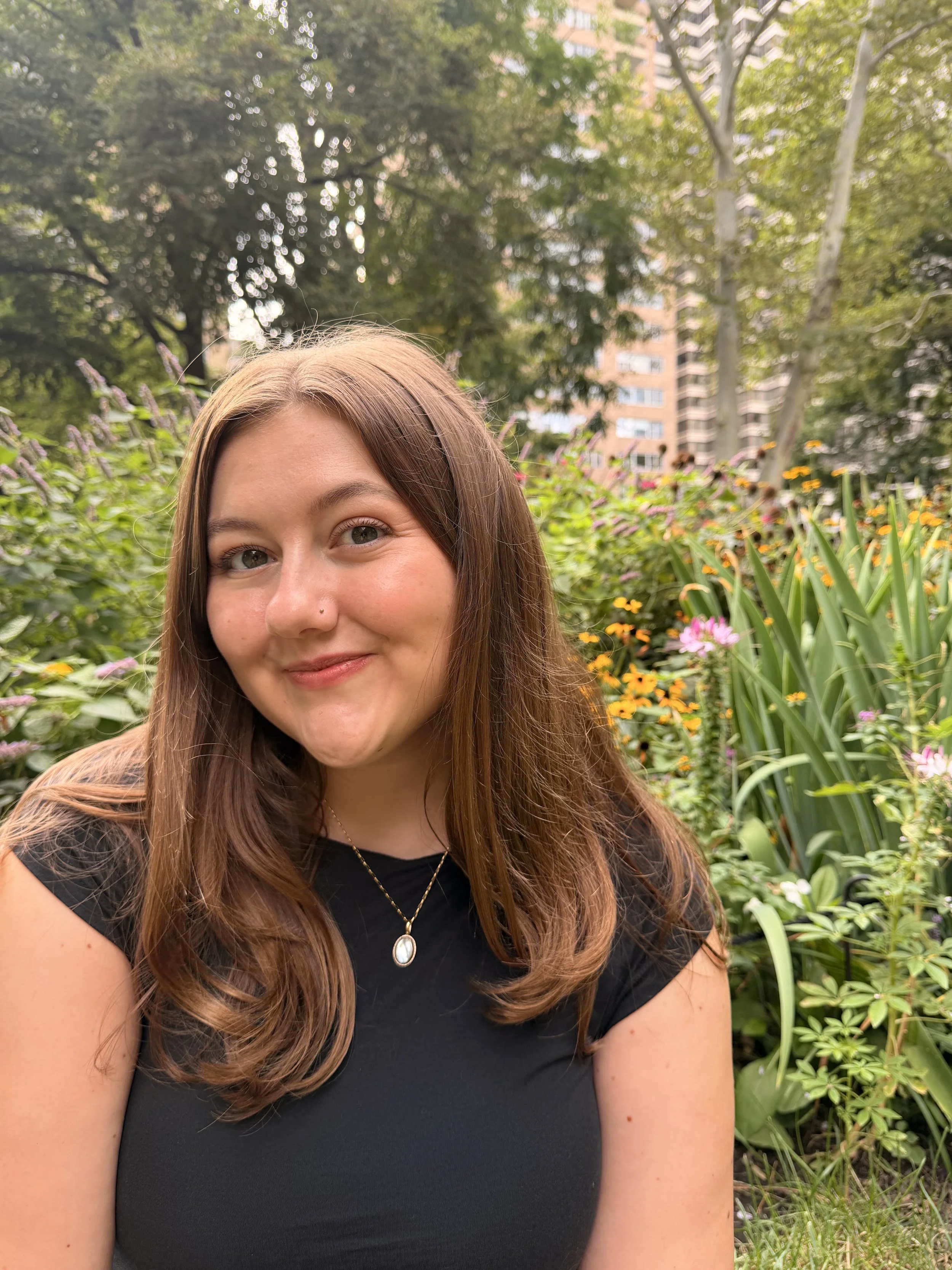 A woman with long brown hair and a black top smiling in a lush garden with colorful flowers and green trees, with buildings in the background.