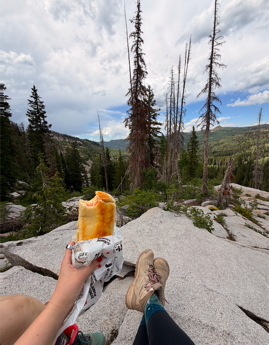 Person holding a partially eaten sandwich or burrito while sitting on a large rock in a mountainous forest area with tall, sparse trees and cloudy sky.