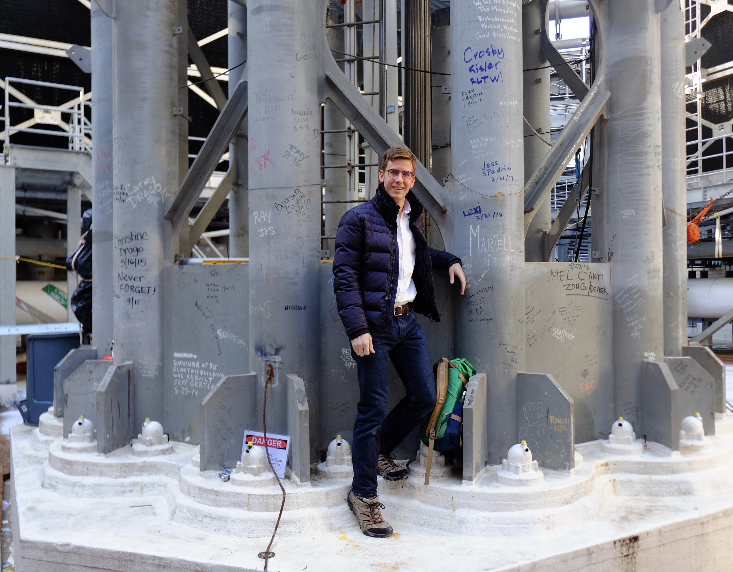 A young man with glasses, a dark jacket, and a backpack standing outdoors in front of large steel beams covered with writings and signatures, at a construction site or monument, with a sign that says 'Danger'. 高  One World Trade Center, 1WTC, NYC