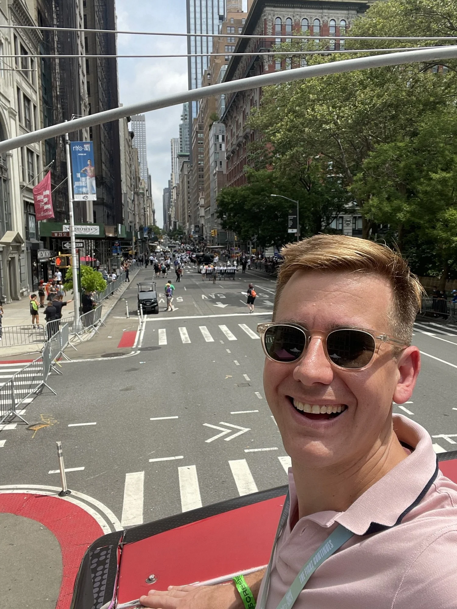 A smiling man wearing sunglasses and a pink polo shirt is standing on a city street, with tall buildings, trees, and people in the background.