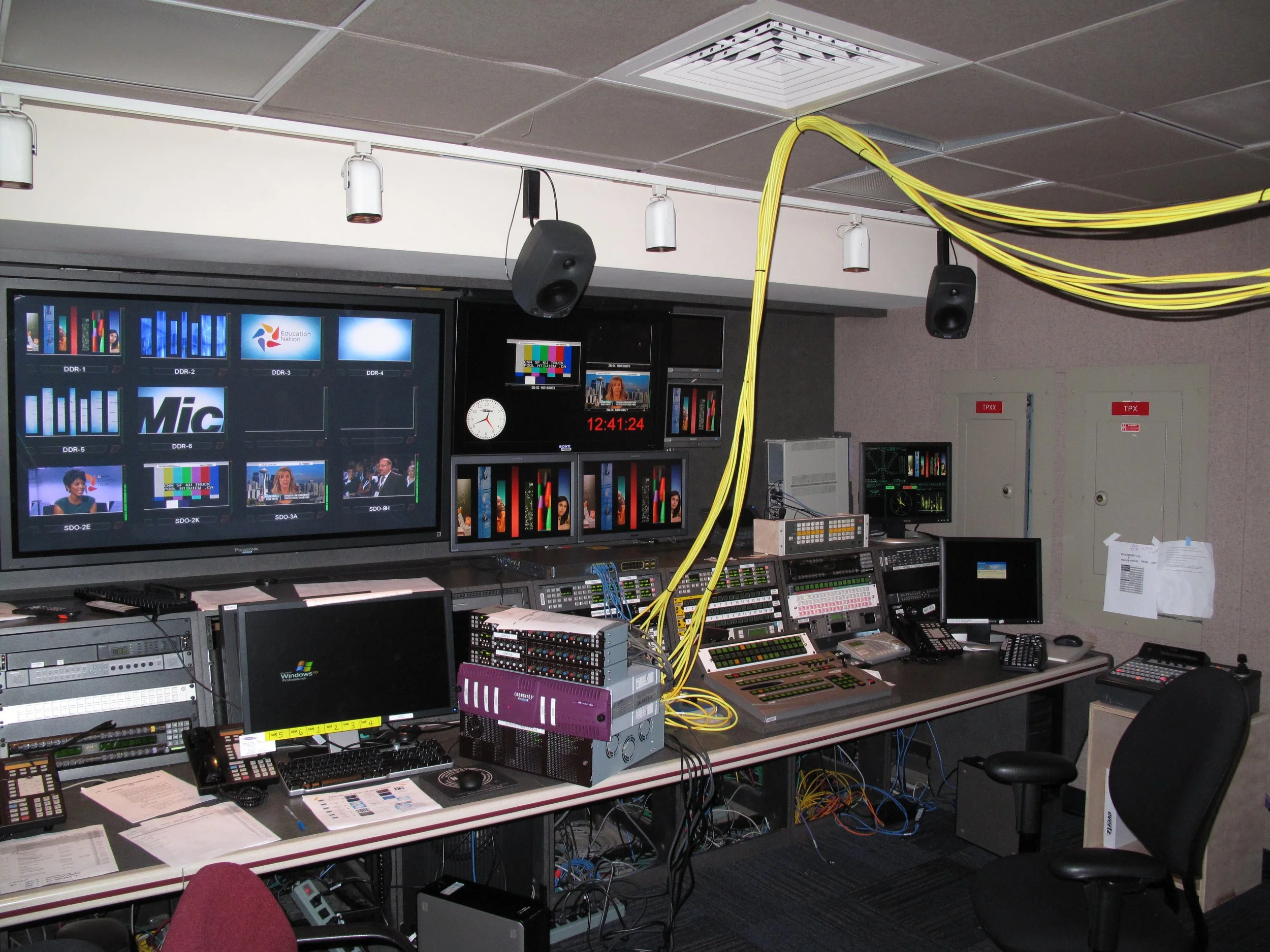 Television control room with multiple monitors displaying news and broadcasts, control panels, computers, and yellow cables hanging from the ceiling.