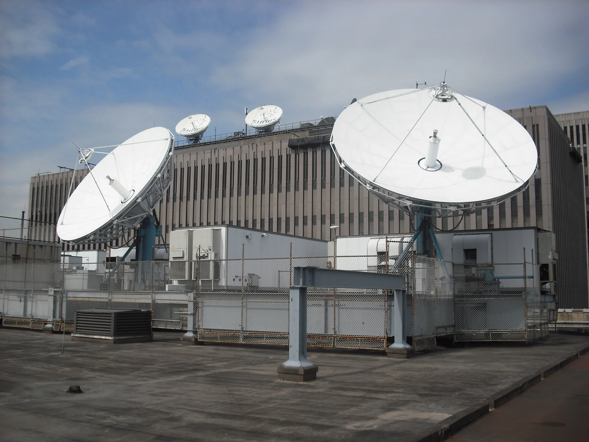 View of rooftop with satellite dishes against a partly cloudy sky, part of a large multi-story building.