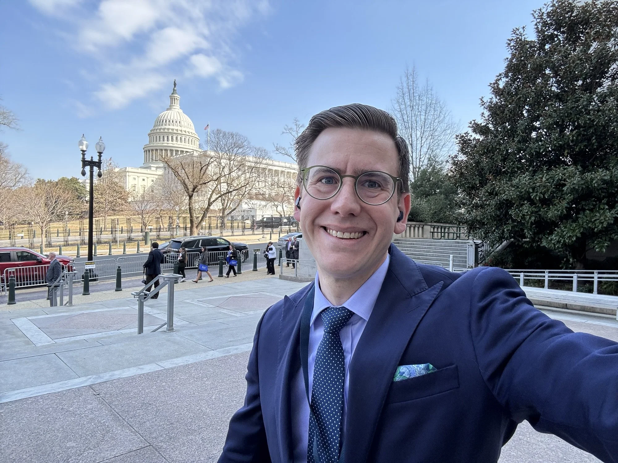 A man in a suit and tie taking a selfie outside the U.S. Capitol building on a clear day with some clouds in the sky.