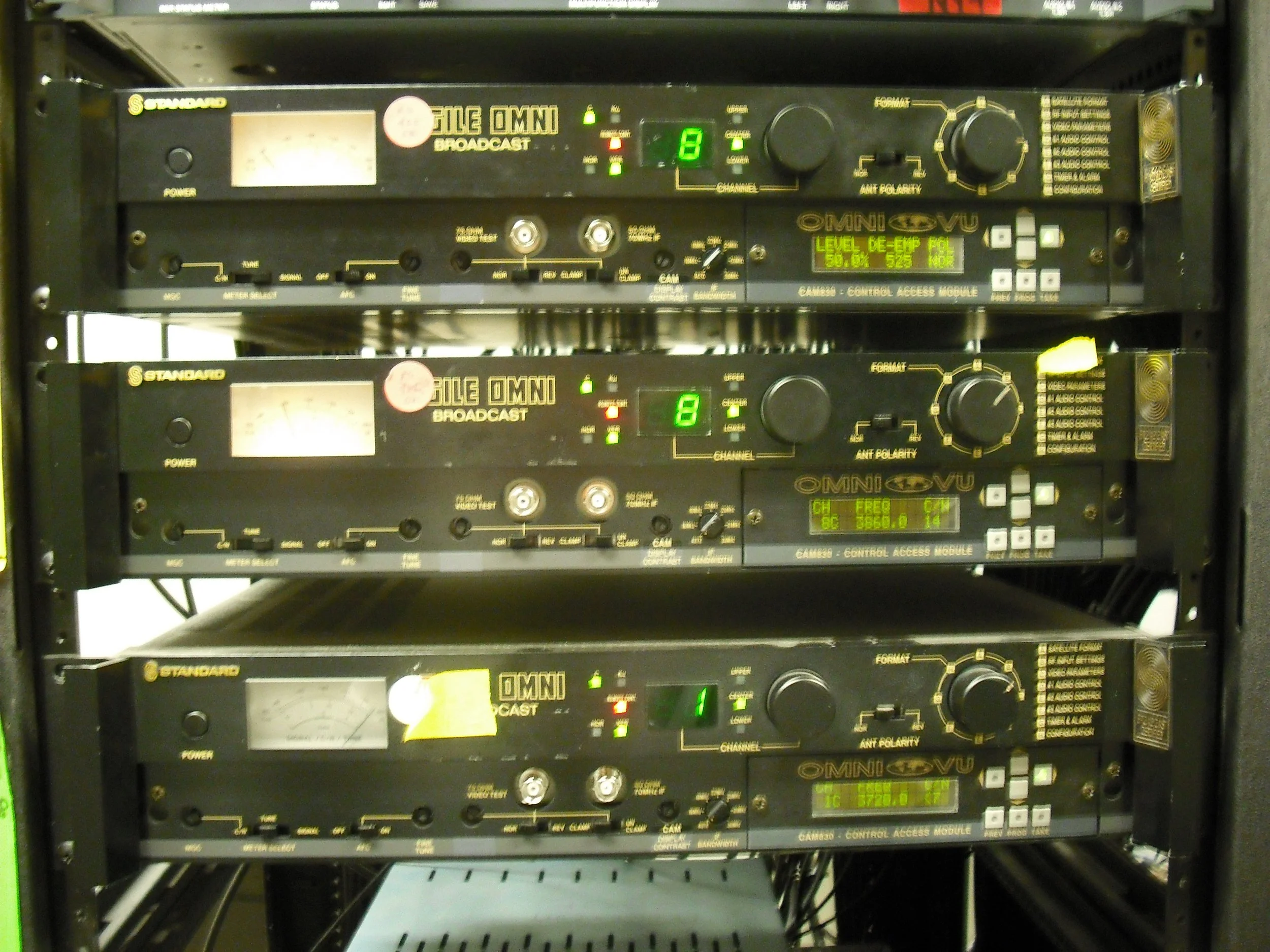 Three racks of broadcast equipment with control modules labeled STANDARD, each featuring various buttons, knobs, and LED displays, mounted on a black metal frame.