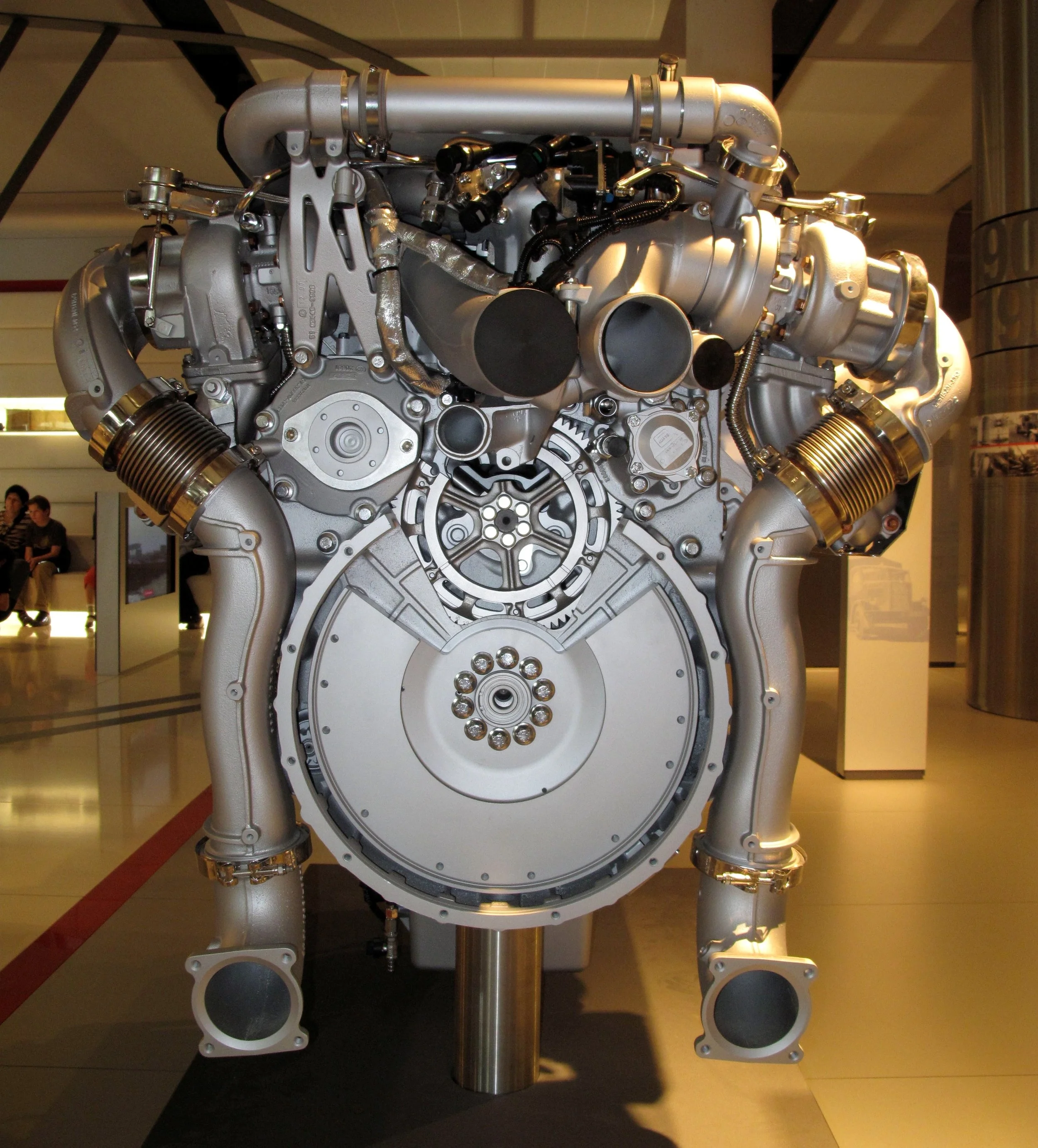 A detailed view of a large, intricate metallic machine component, possibly an engine or mechanical system, displayed indoors with surrounding people.