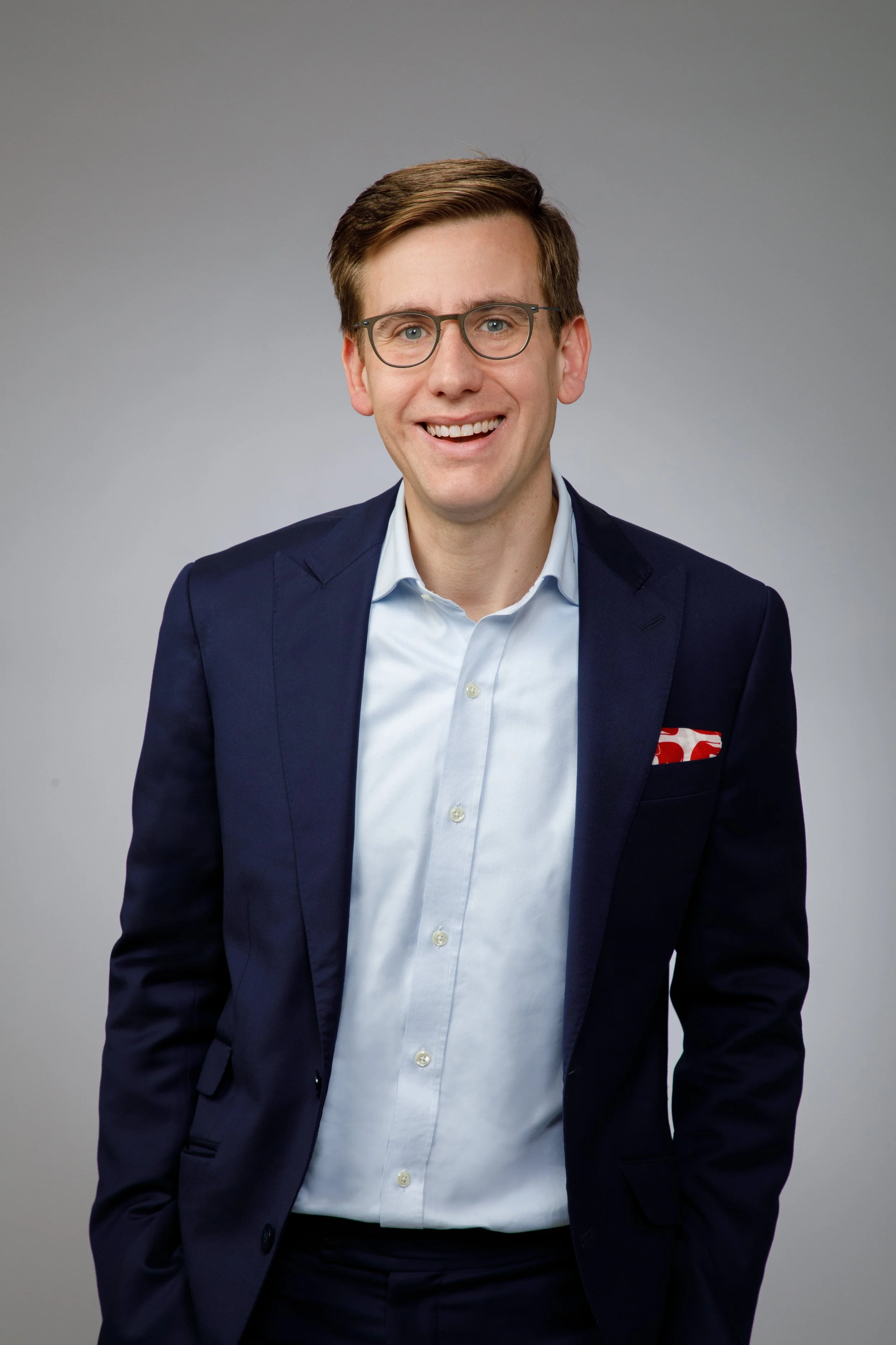 A man wearing a navy blazer, light blue dress shirt, and glasses, smiling at the camera against a plain gray background.