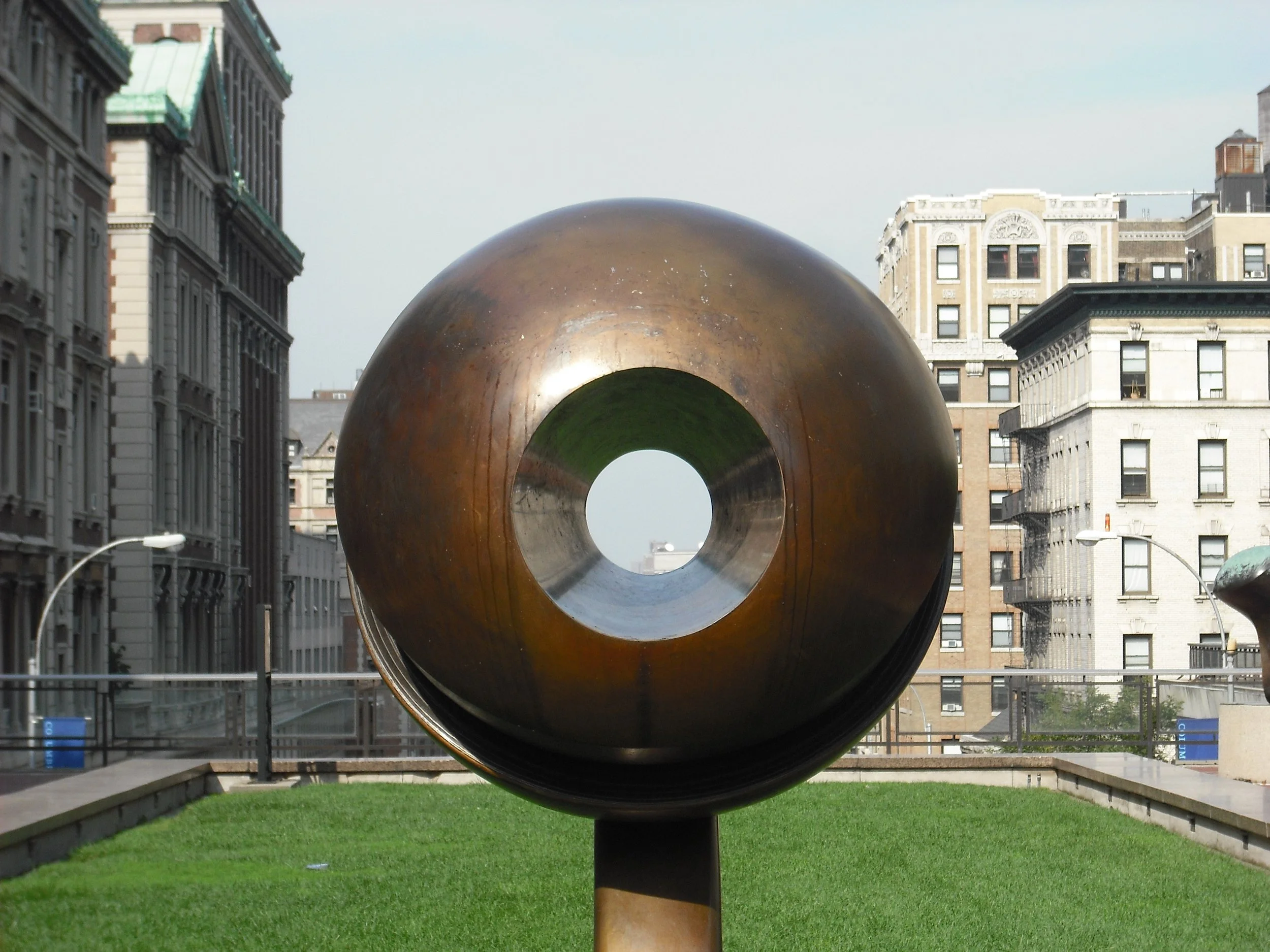 A large circular metallic sculpture situated in an urban park, with surrounding buildings and a grassy area. Columbia University, Morningside Heights, New York City, NYC