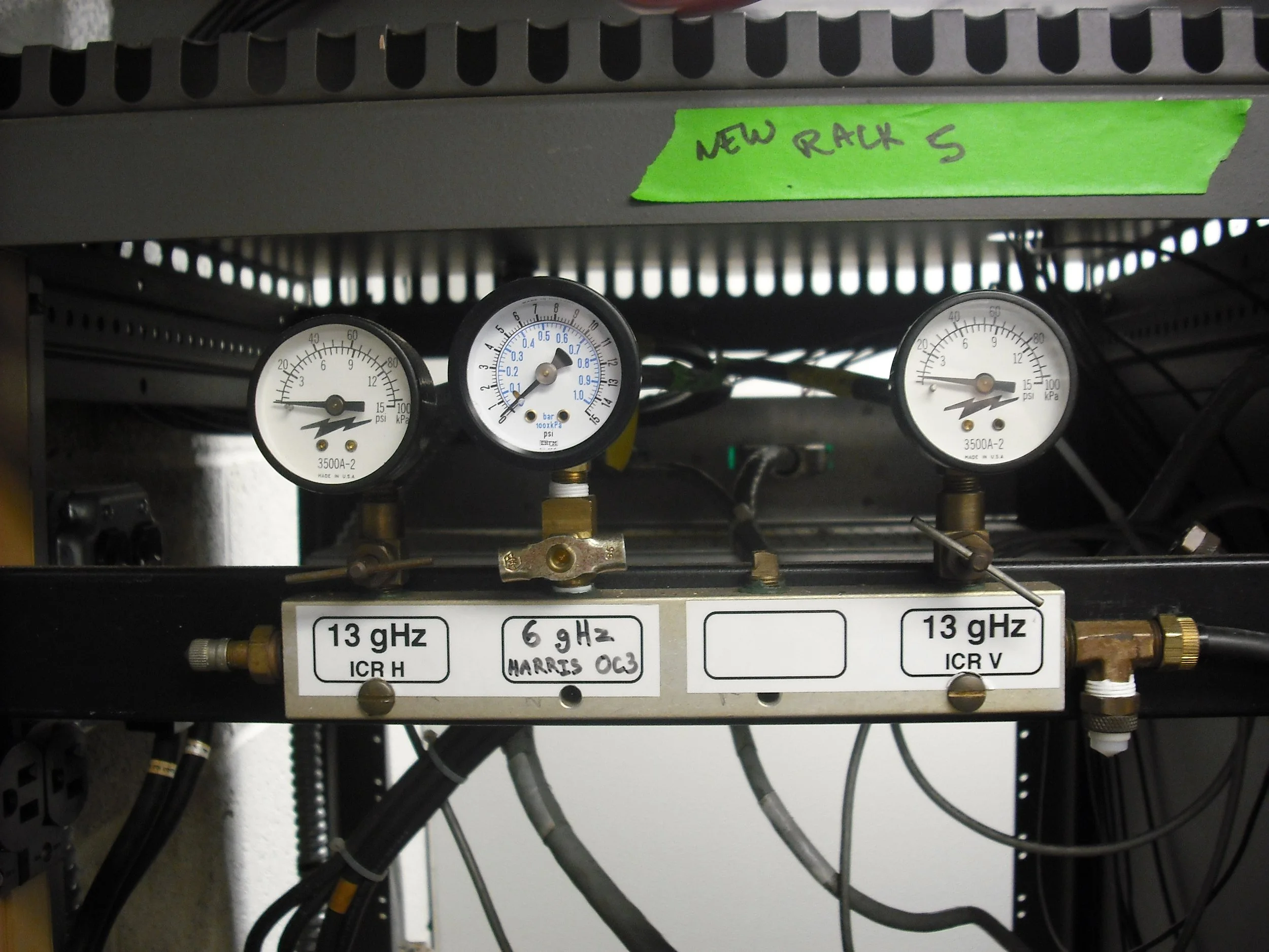 Equipment with three pressure gauges and labels indicating frequencies of 13 GHz and 6.9 GHz, with a green tape label reading 'NEW RACK S'.