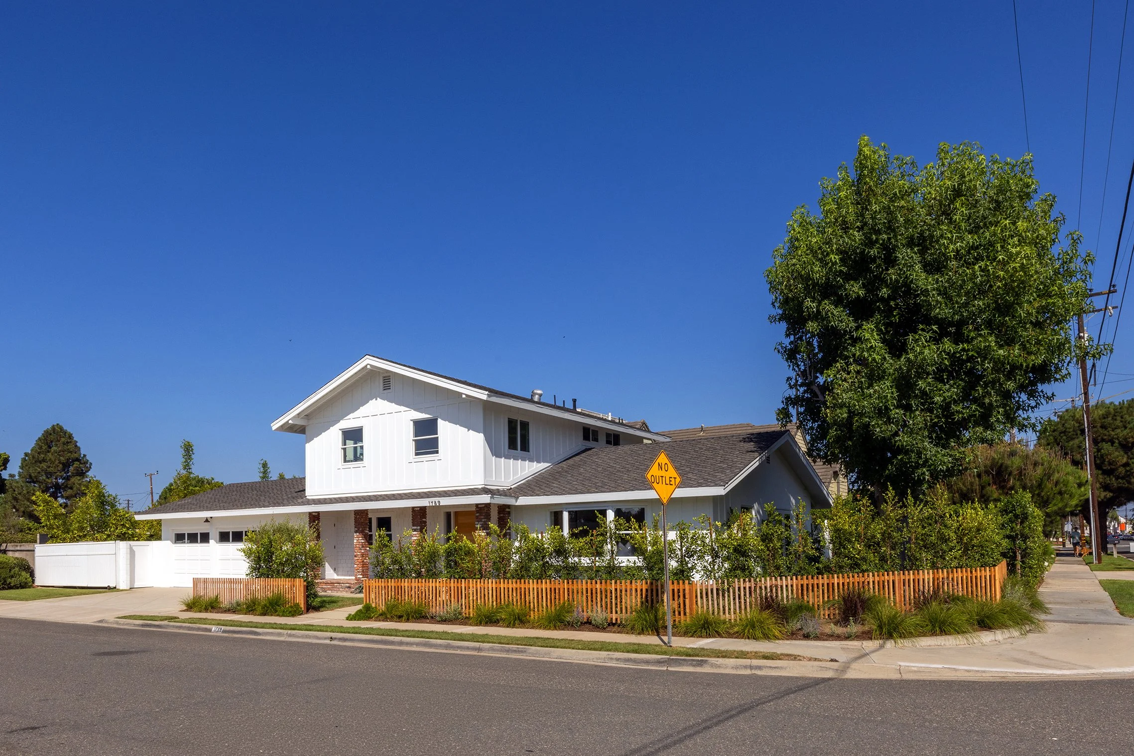 White two-story house with a gabled roof, brick chimney, and a wooden fence, situated on a street corner with a 'No Outlet' sign in the yard, surrounded by trees and clear blue sky.
