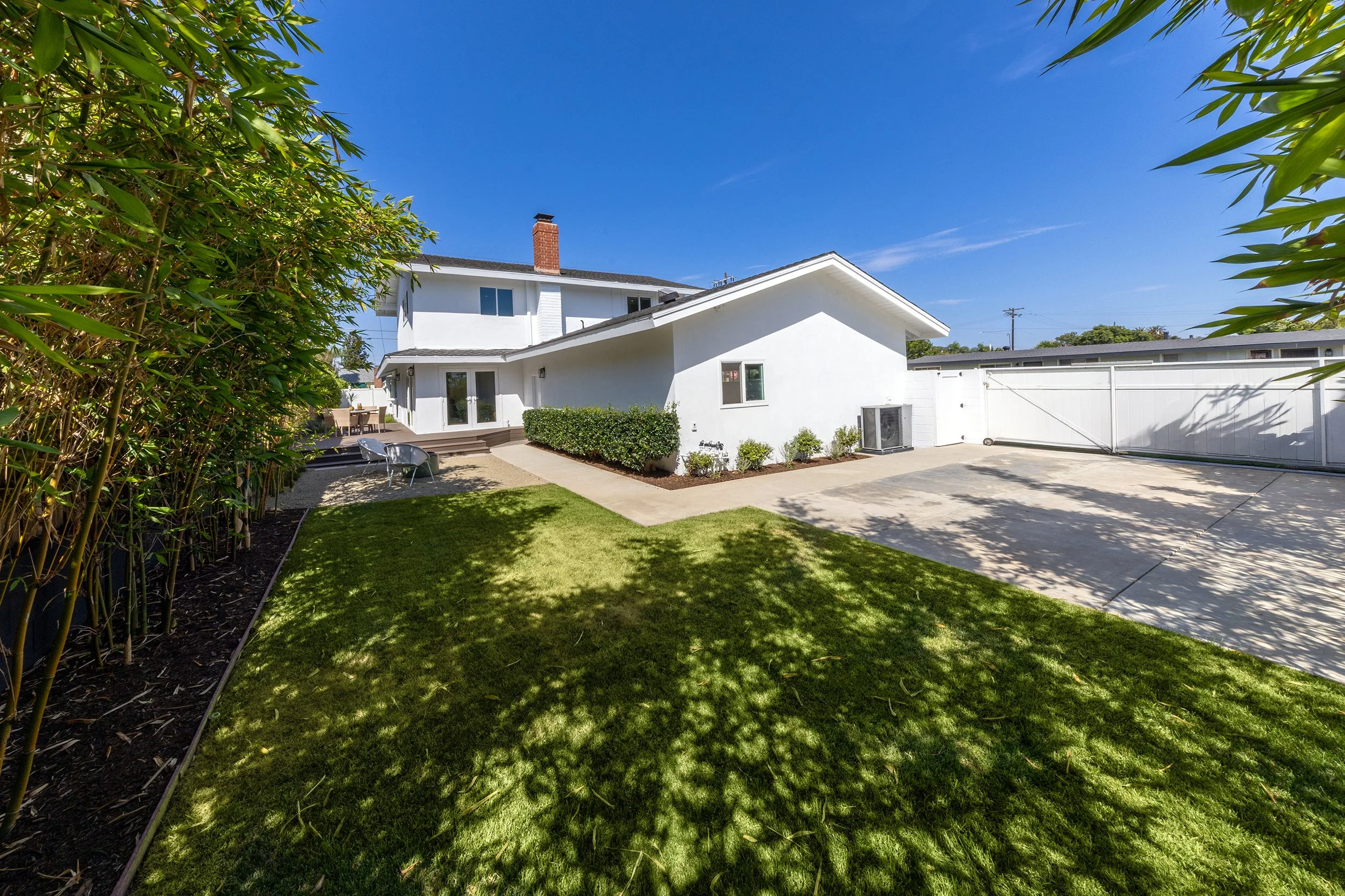 Backyard with a well-maintained lawn, patio furniture, and a white house with a sloped roof under a bright blue sky.