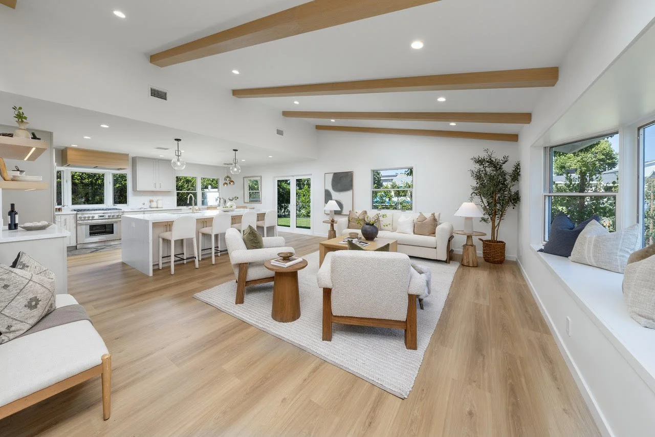Spacious open-concept living room and kitchen with white walls and wooden accents, featuring large windows, light wood flooring, white sofas, armchairs, and modern decor.