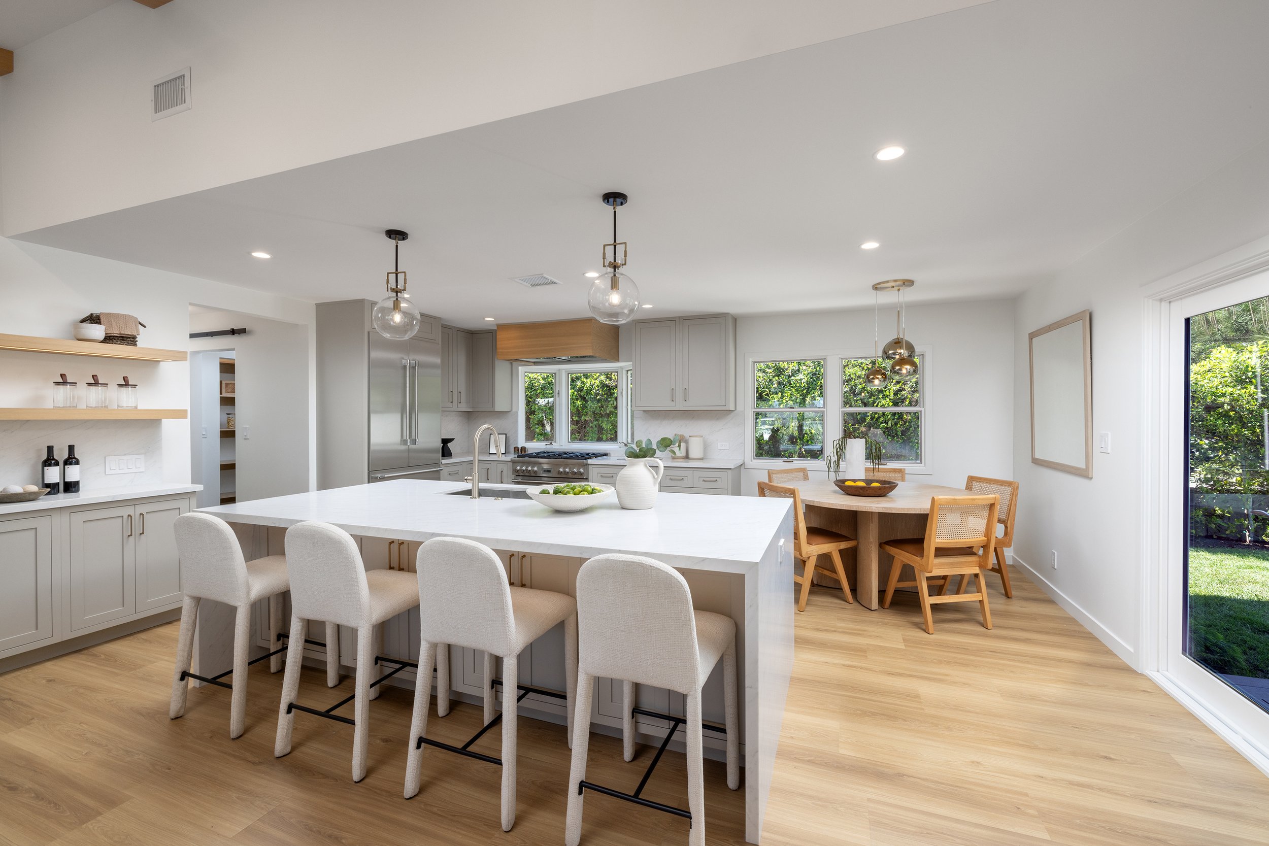 Modern, open-concept kitchen with white cabinetry, large island with seating, pendant and recessed lighting, light wood flooring, and large windows with greenery outside.
