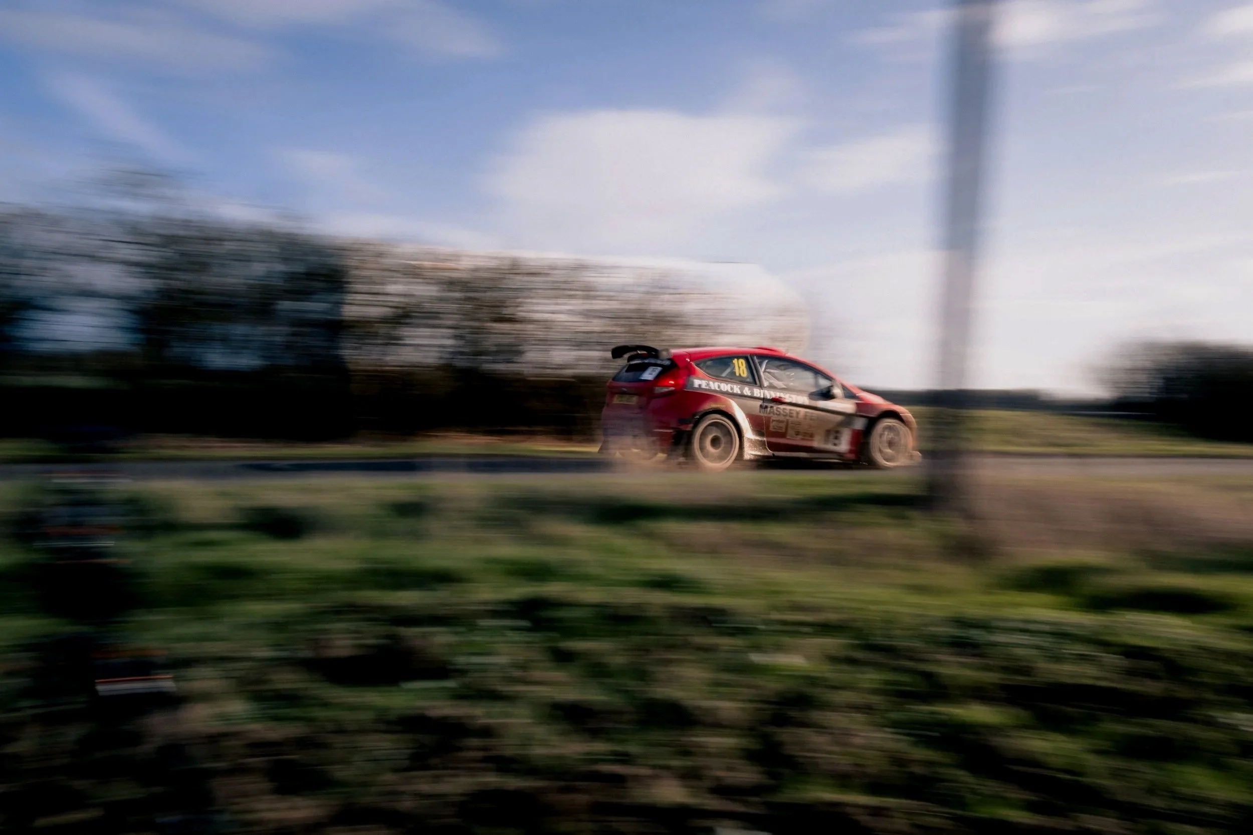 A red rally car with racing decals speeding on a road, creating motion blur with a background of trees and a partly cloudy sky.