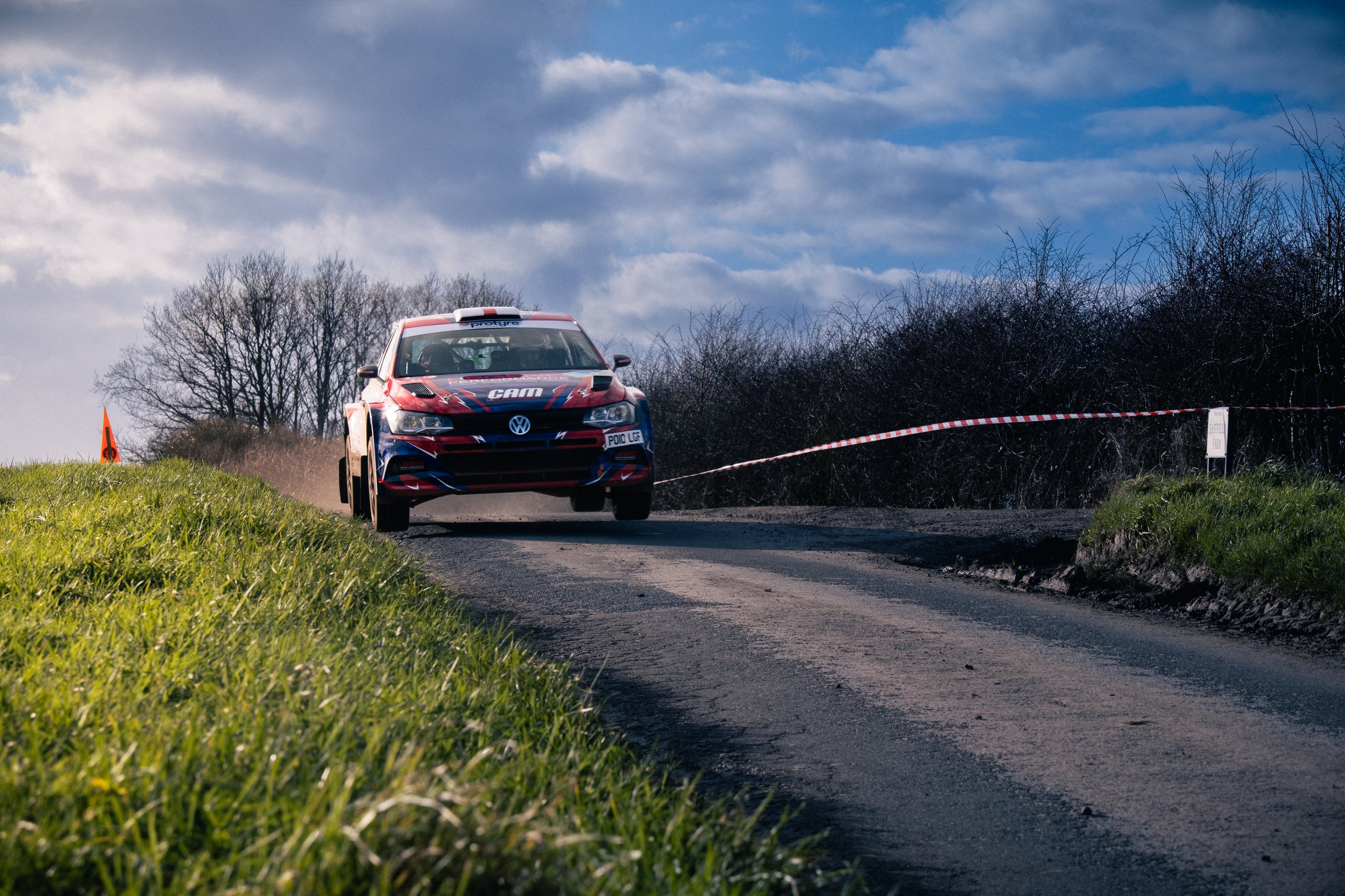 A rally car driving on an Asphalt road at East Riding Stages in a rural area with trees and clouds in the background.