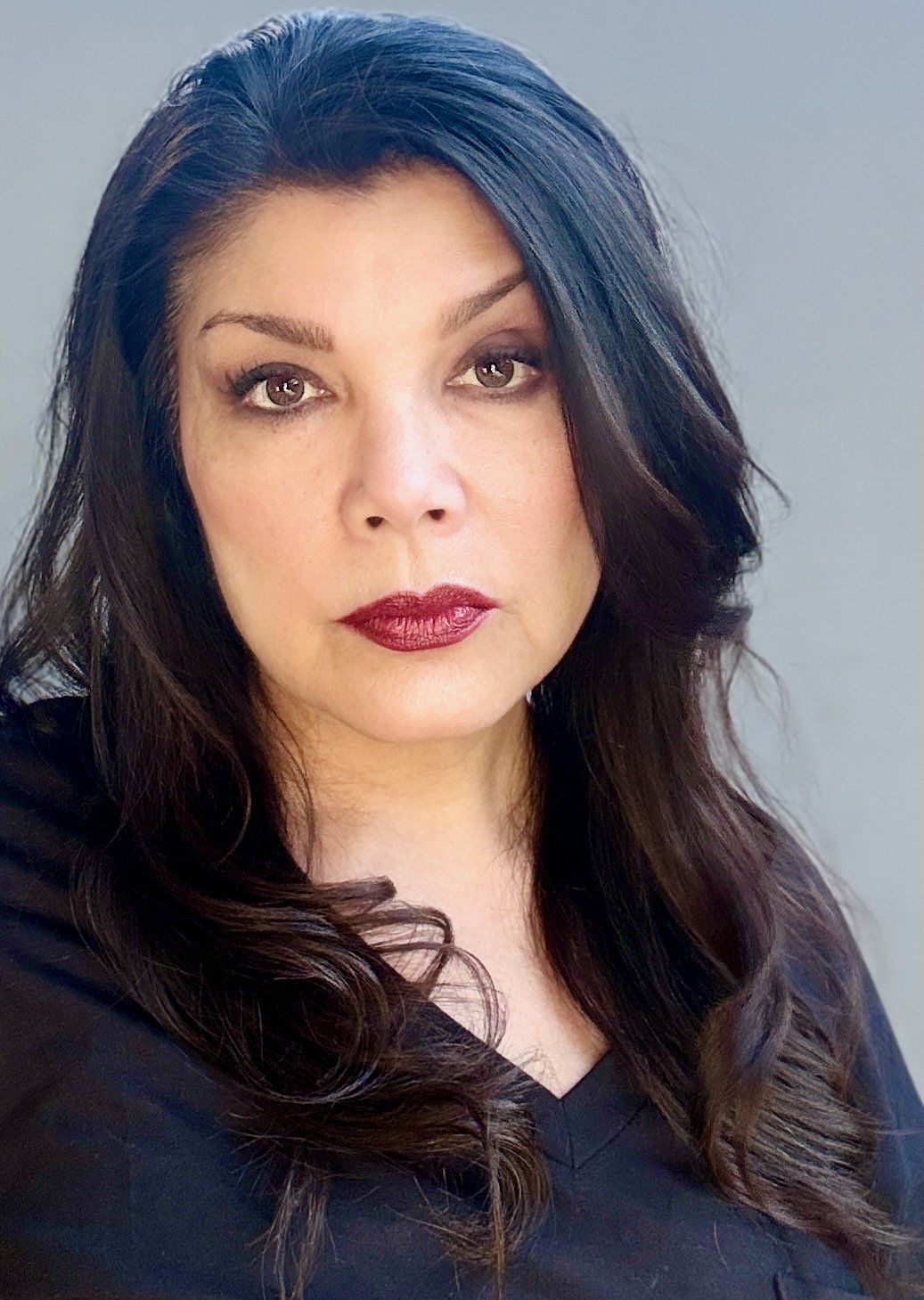 A woman with dark brown, wavy hair and makeup, wearing a black top, looking at the camera.