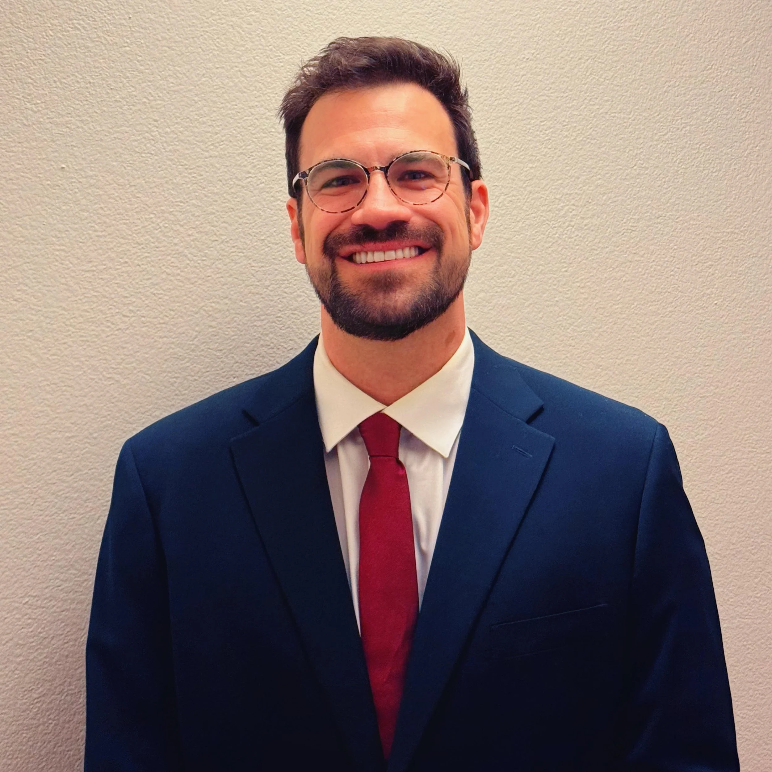 A man with glasses smiling, wearing a suit and tie, standing against a plain beige wall.