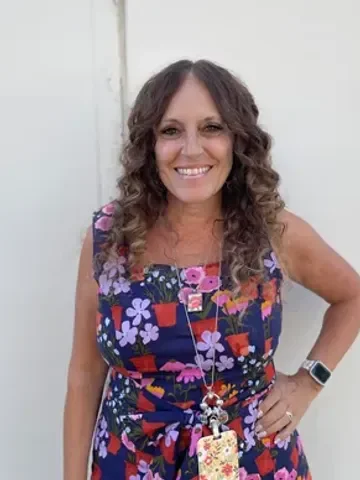 Woman with curly brown hair wearing a colorful floral dress, smiling, standing against a plain wall.