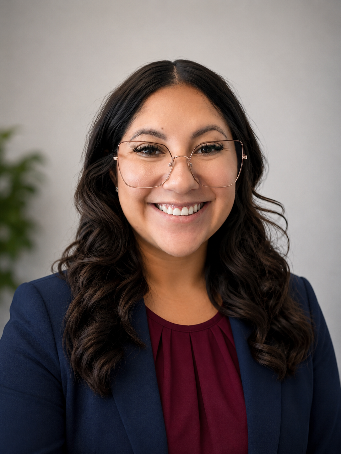 Professional woman with glasses and dark, wavy hair, smiling, wearing a navy blazer and maroon top, indoors with neutral background and a blurred green plant.