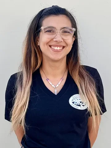 Smiling woman with long, wavy hair, wearing glasses, a black polo shirt with a logo, and a beaded necklace.