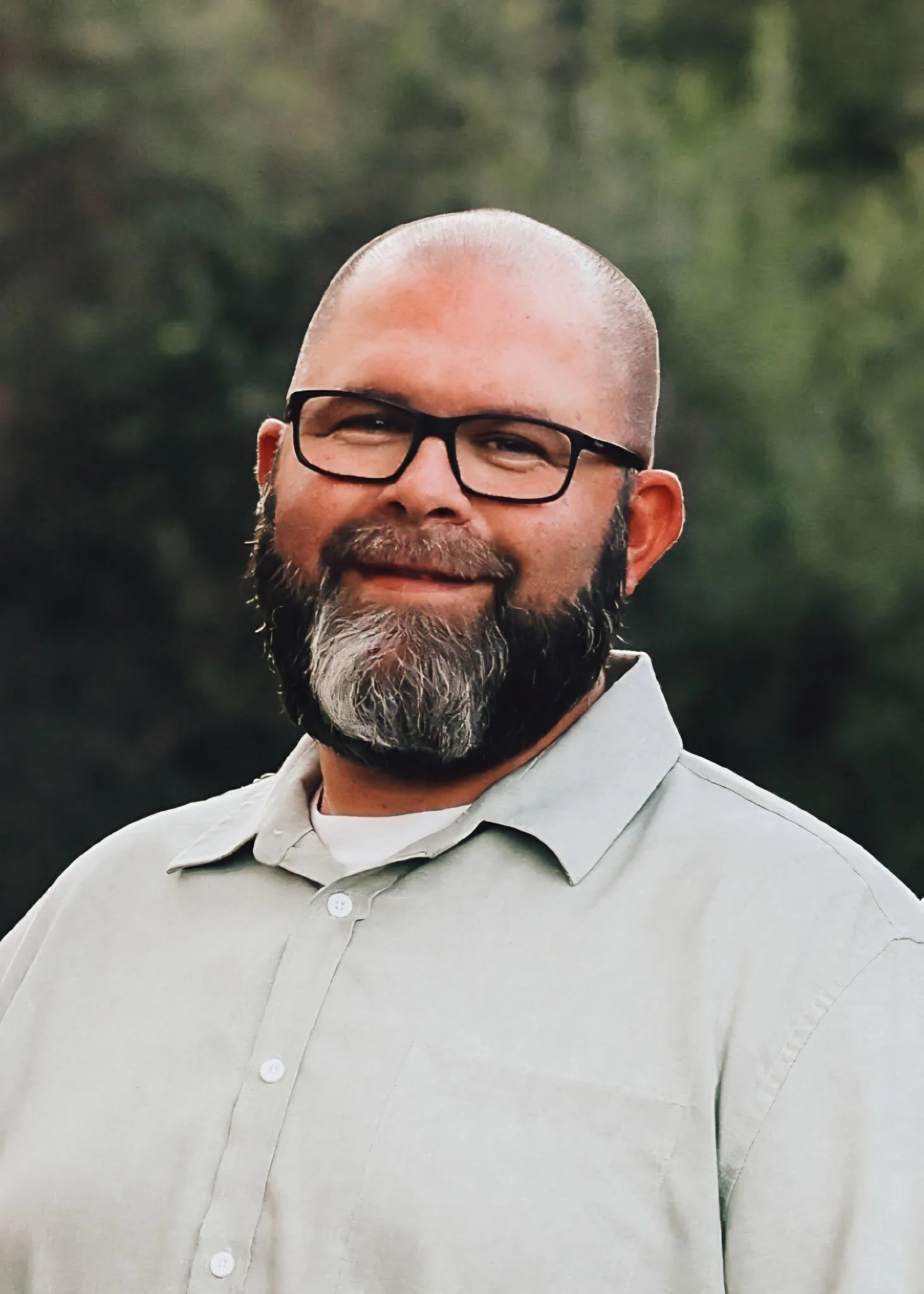 A man with a bald head, glasses, and a beard, wearing a light-colored button-up shirt, smiling outdoors with a blurry green background.