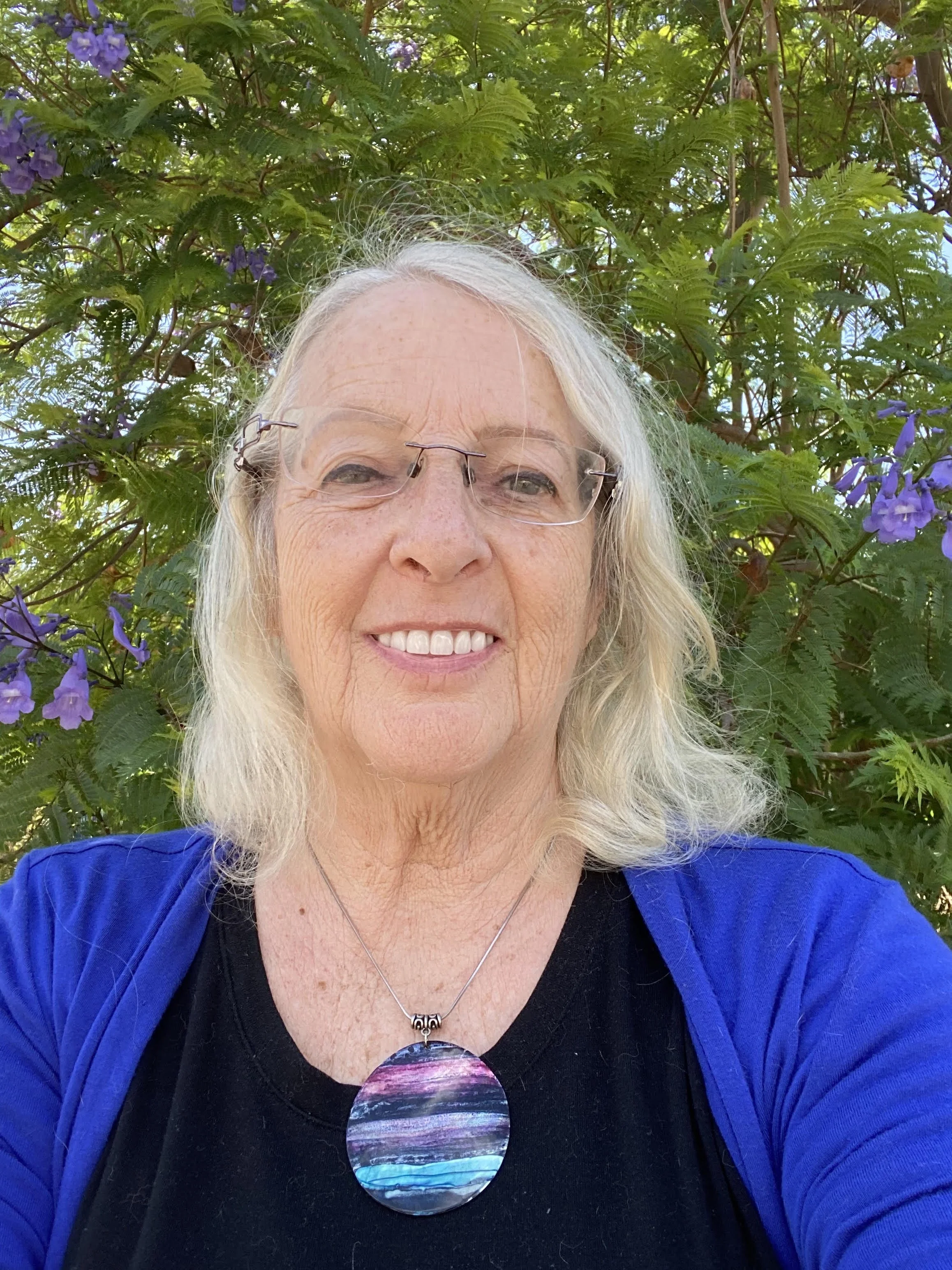 A smiling woman with gray hair wearing glasses, a black top, and a blue jacket, standing outdoors in front of a leafy green plant with purple flowers.