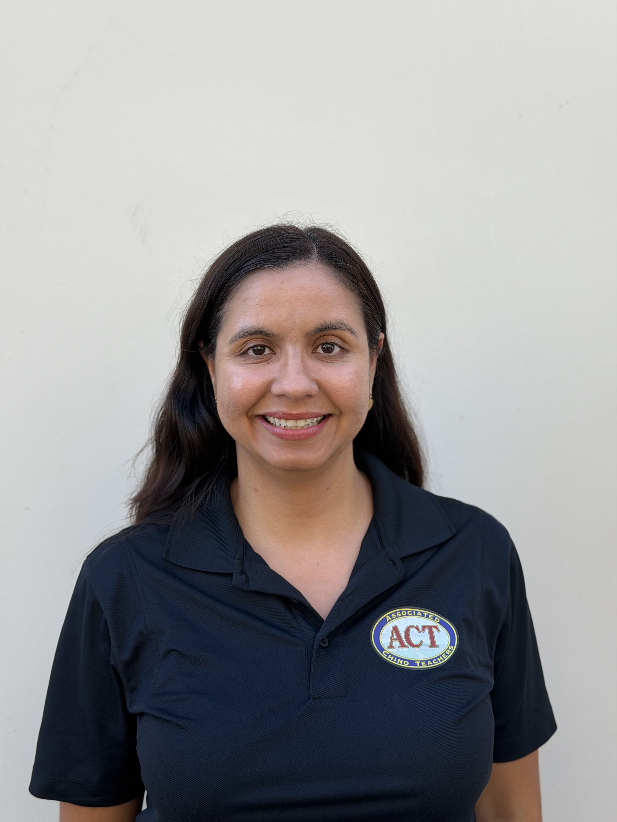 A woman with long dark hair smiling, wearing a navy blue polo shirt with a badge that says 'ACT' and 'Chino Teachers' in front of a plain white wall.