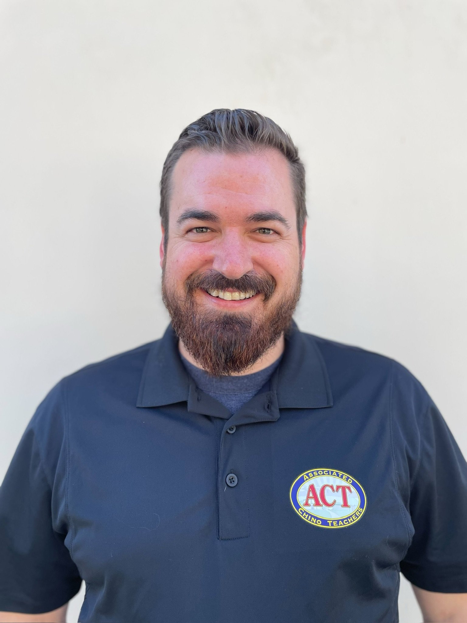 A man wearing a black polo shirt with a logo that reads 'ACT' and 'Chino Teachers' smiling in front of a plain light-colored background.