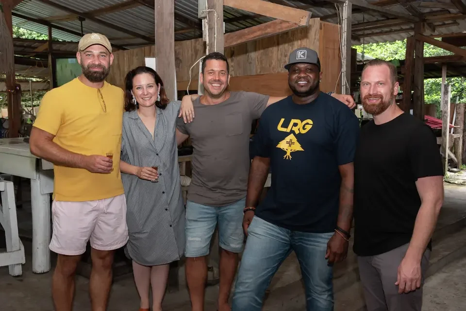 Five friends standing together indoors, smiling and enjoying drinks, with a rustic wooden structure in the background.