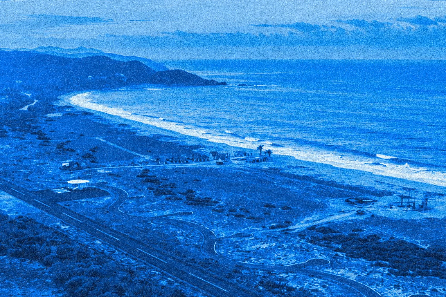 An aerial view of a coast with a curving shoreline, waves breaking on the beach, and a hilly landscape in the background. There are roads and a few scattered structures near the beach.
