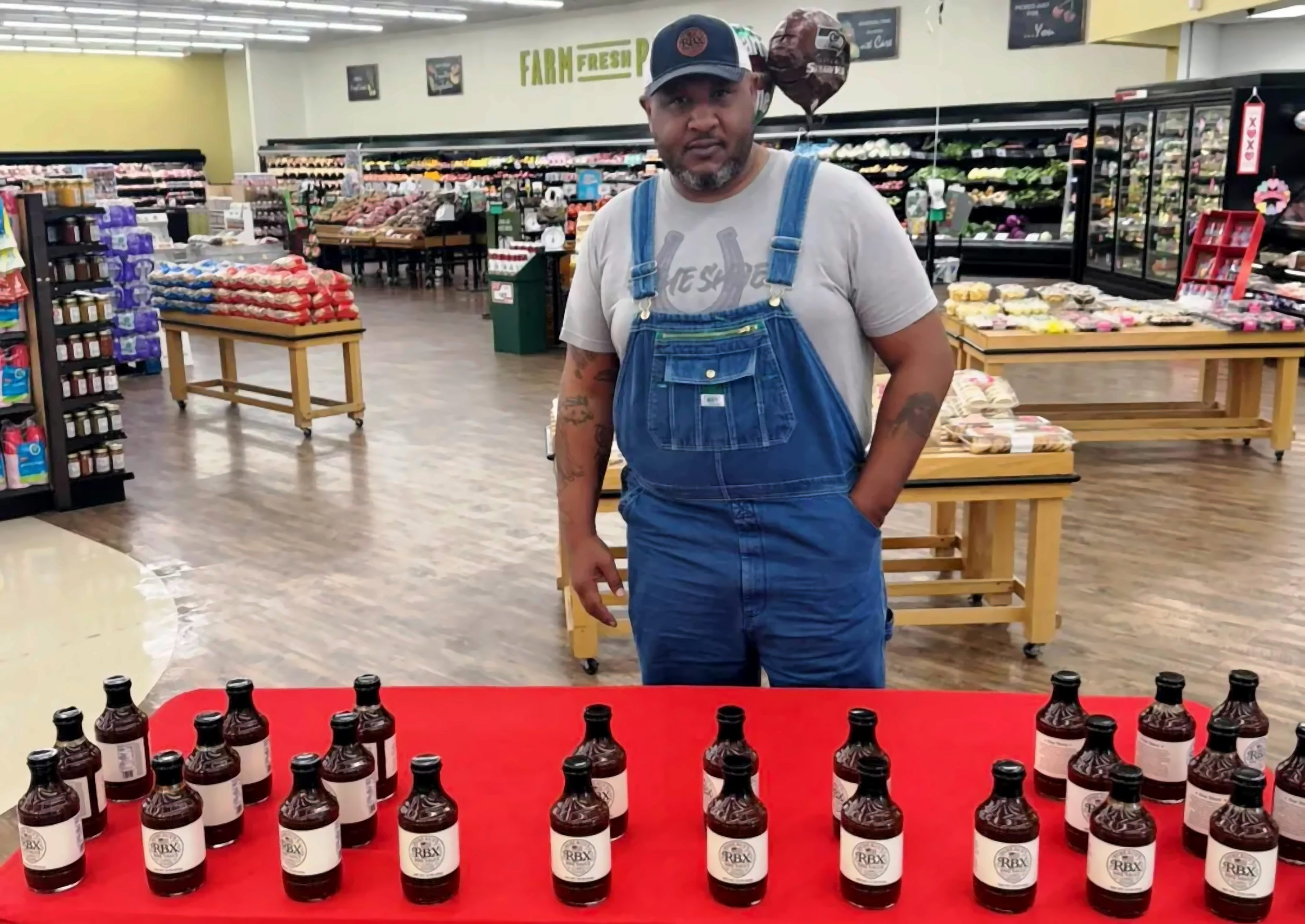 Man in jean overalls and a cap standing inside a grocery store, with displays of sauces or condiments on a red table in front of him.